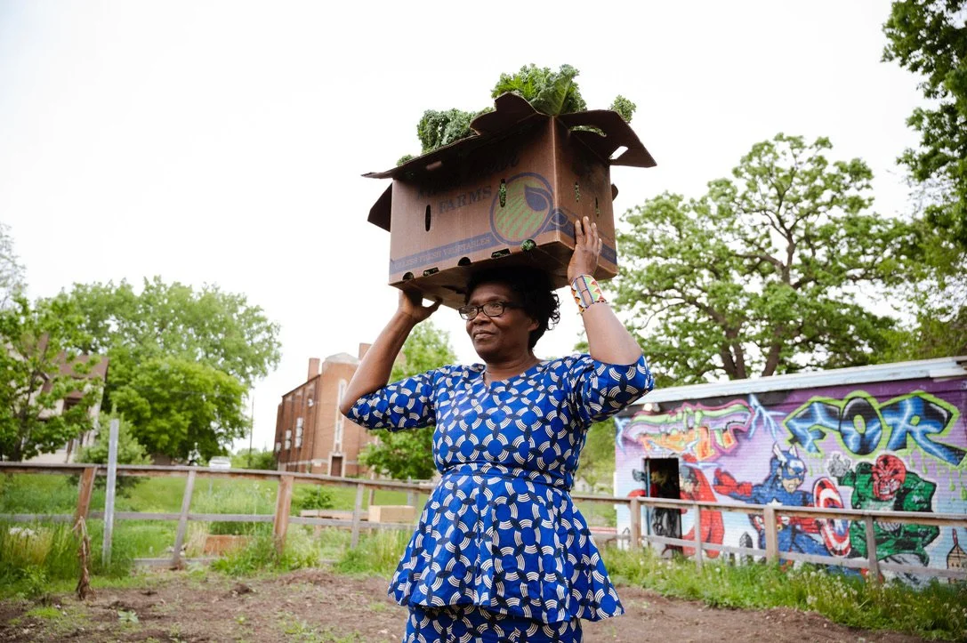 An African woman carrying a cardboard box with vegetables on her head, standing outdoors in an urban garden with trees and a graffiti-covered wall in the background.