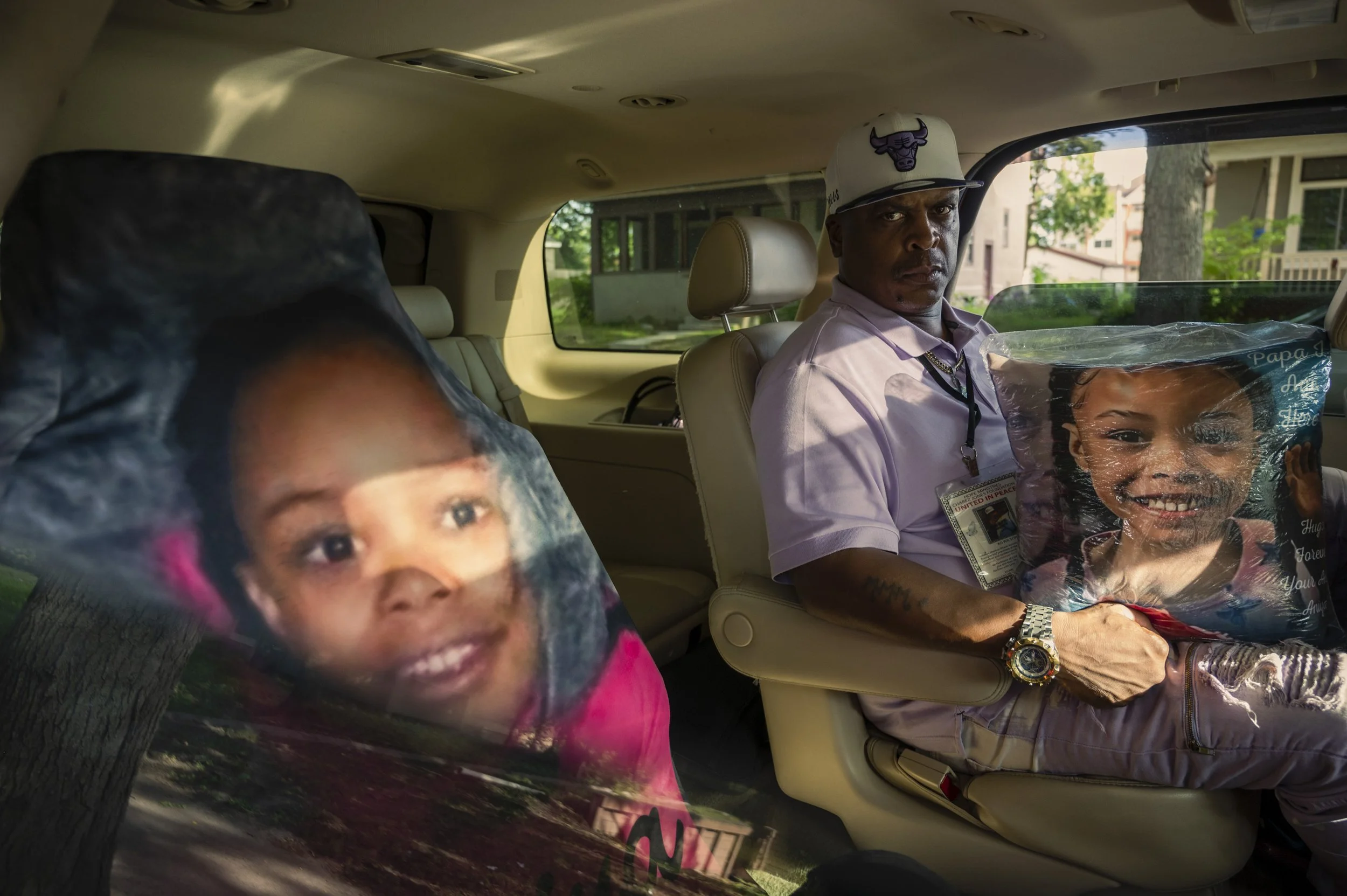 A man in a white shirt and a Chicago Bulls cap sitting in the backseat of a car, holding a pillow with a child's face on it. The pillow shows a smiling young girl. The man has a serious expression and is wearing an ID badge.