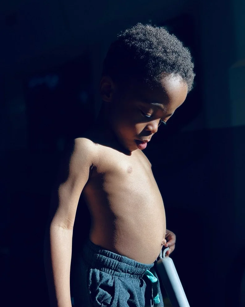 A young Black boy and short curly hair, looking down at his stomach in a side profile view, illuminated by hard natural light.