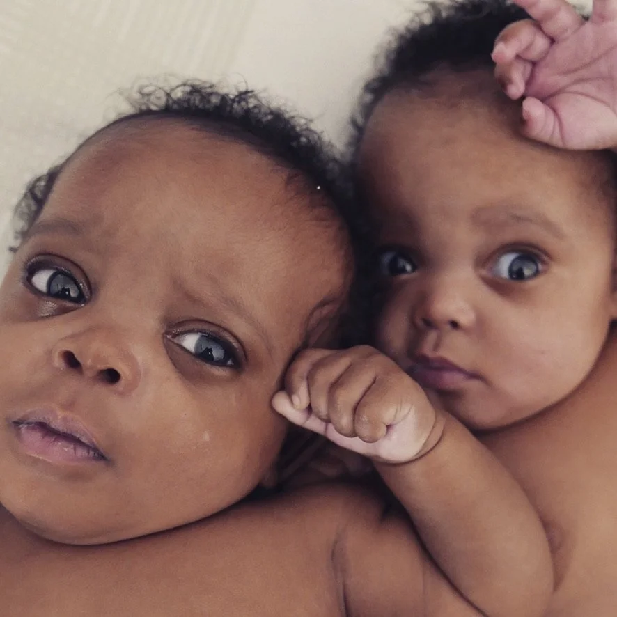 Two young babies with curly hair lying close together, looking towards the camera, one with a hand near their face.