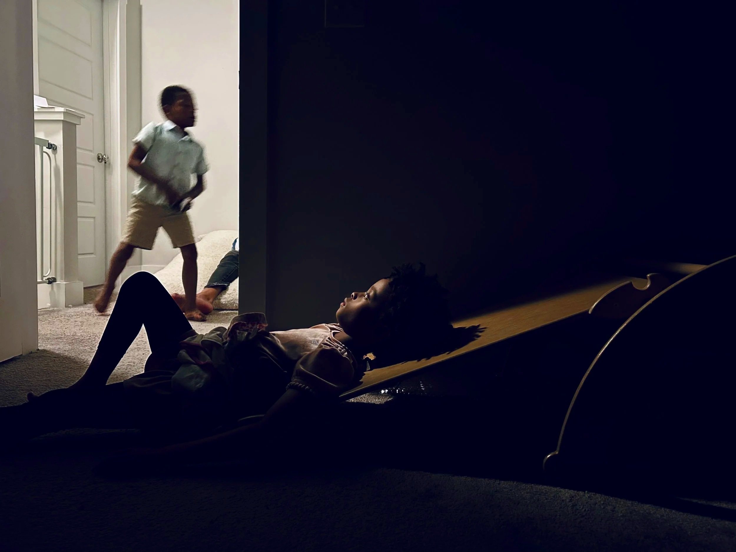 A child lying on the floor with her eyes gazing towards the ceiling in a dimly lit room, while a young boy is running or jumping past her in a brightly lit hallway.