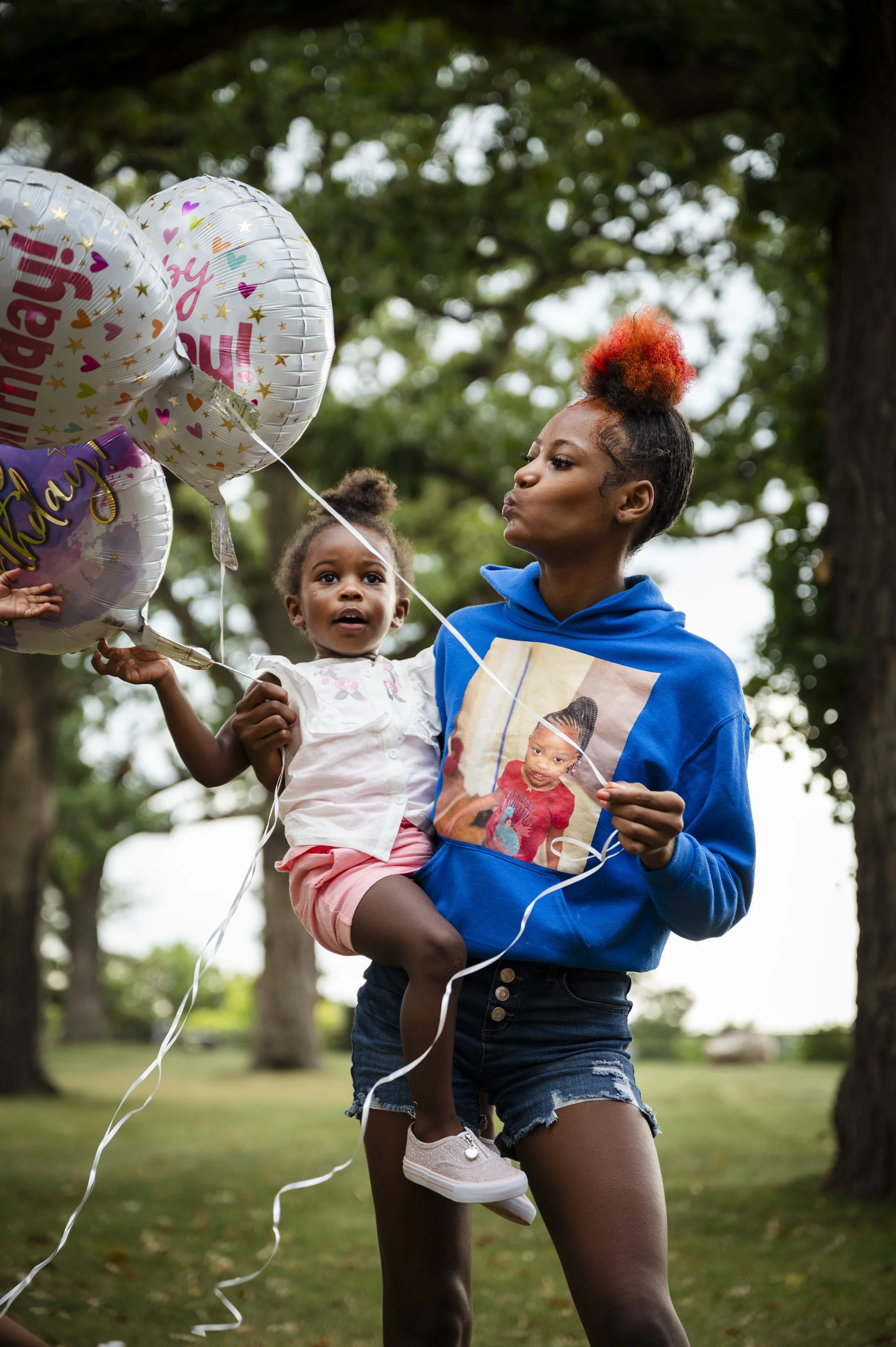 A woman holding a young girl in a park with trees in the background. The woman is wearing a blue hoodie with a photo of a child on it and has a colorful, fluffy updo. The girl is holding balloons with celebratory messages, and she is wearing a white 