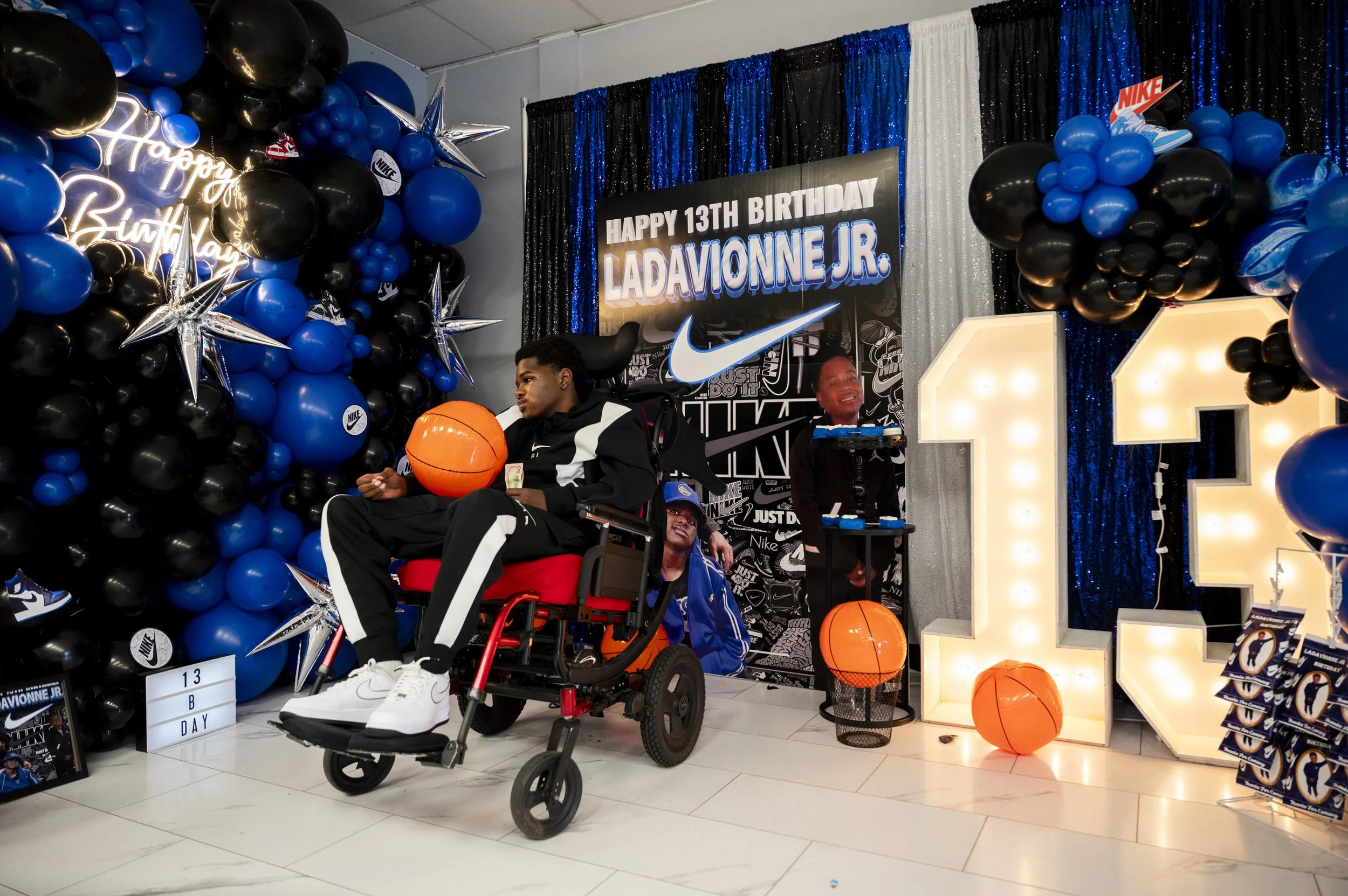 A boy in a wheelchair celebrates his 13th birthday with a sports-themed party, decorated with blue, black, and white balloons, large illuminated number 13, a basketball, Nike banners, and a sign that reads 'Happy 13th Birthday Ladavonne Jr.'