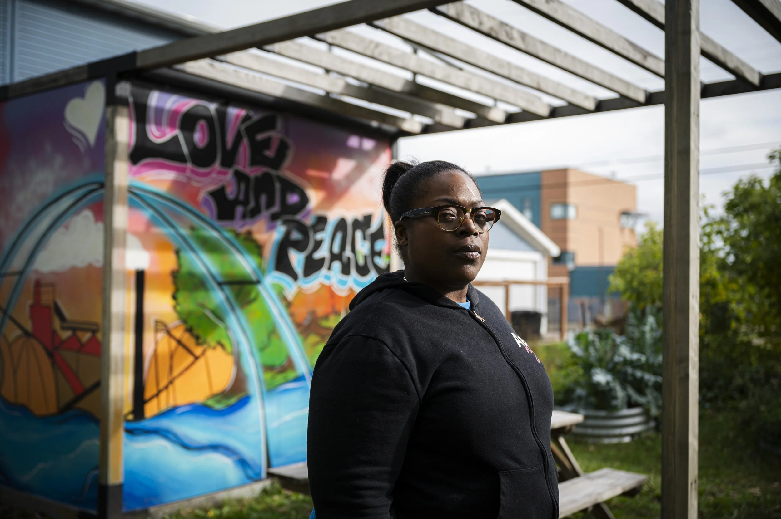 A woman with glasses standing outdoors in front of a colorful mural that says "Love and Peace" with a globe and rainbow design. The mural was made to commemorate her son who died from a shooting.