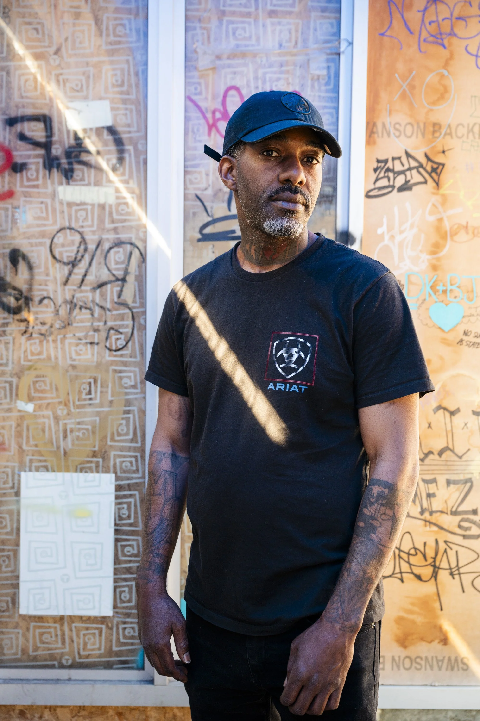 A man wearing a black cap and black T-shirt standing in front of a graffiti-covered wall.