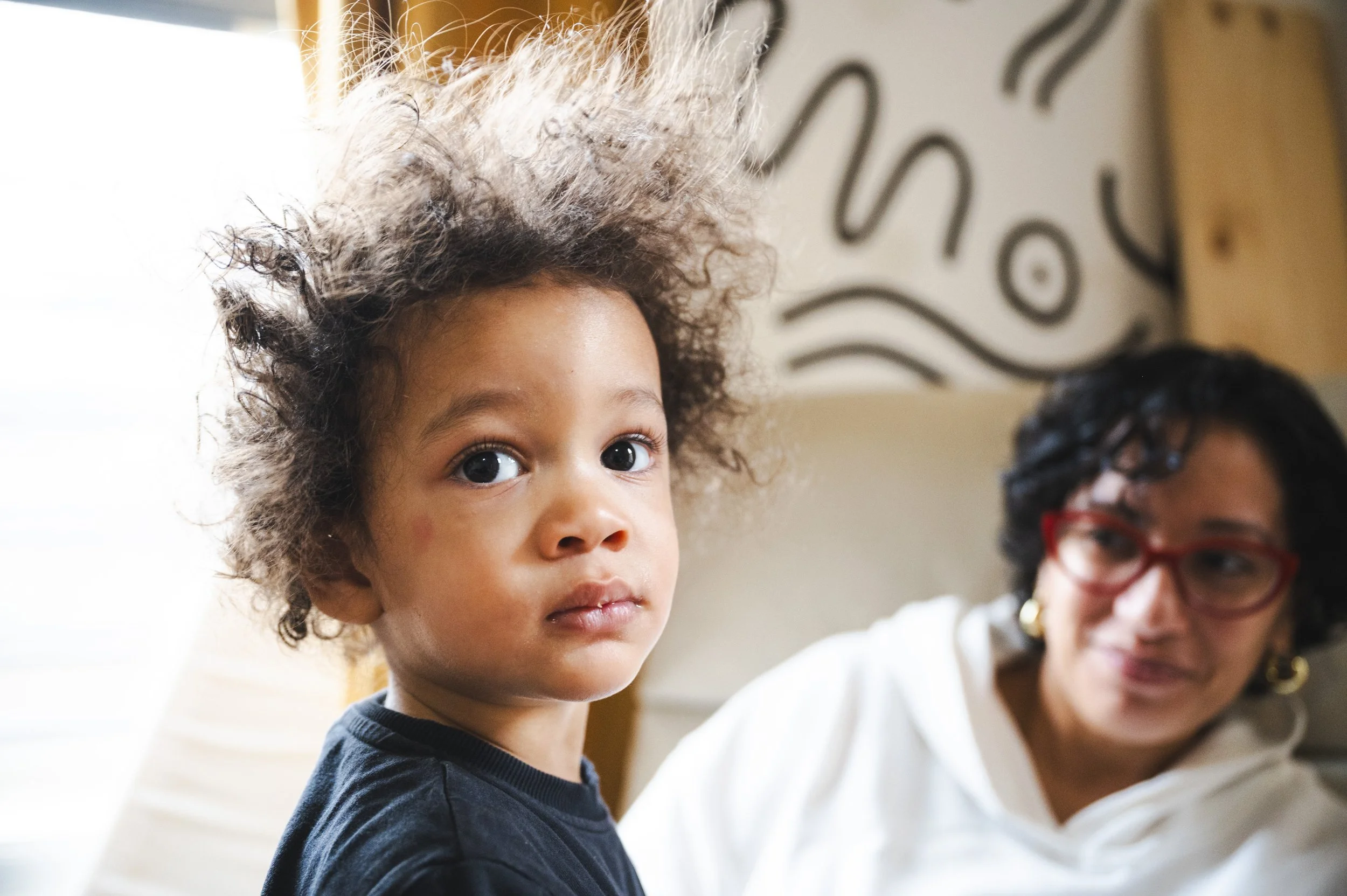 A young Latino child with curly, messy hair looking directly at the camera, with a woman (his mother) with glasses in the background looking at the child.