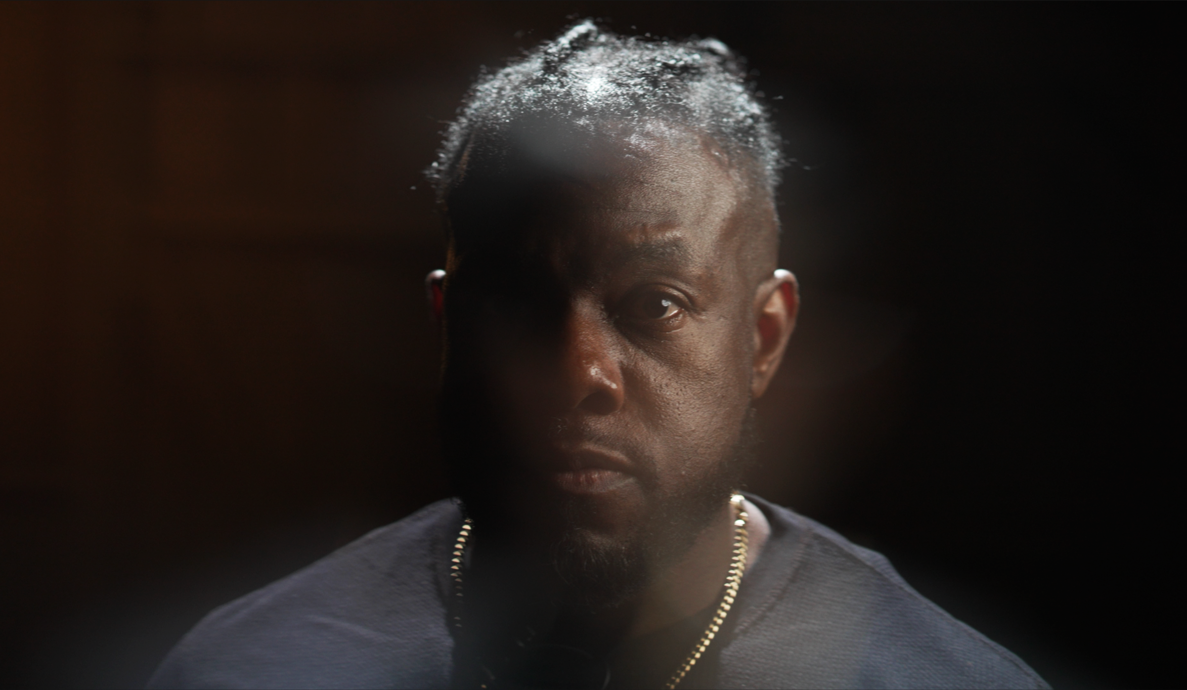 A portrait of a Black man with dreads, looking directly at the camera against a dark background.