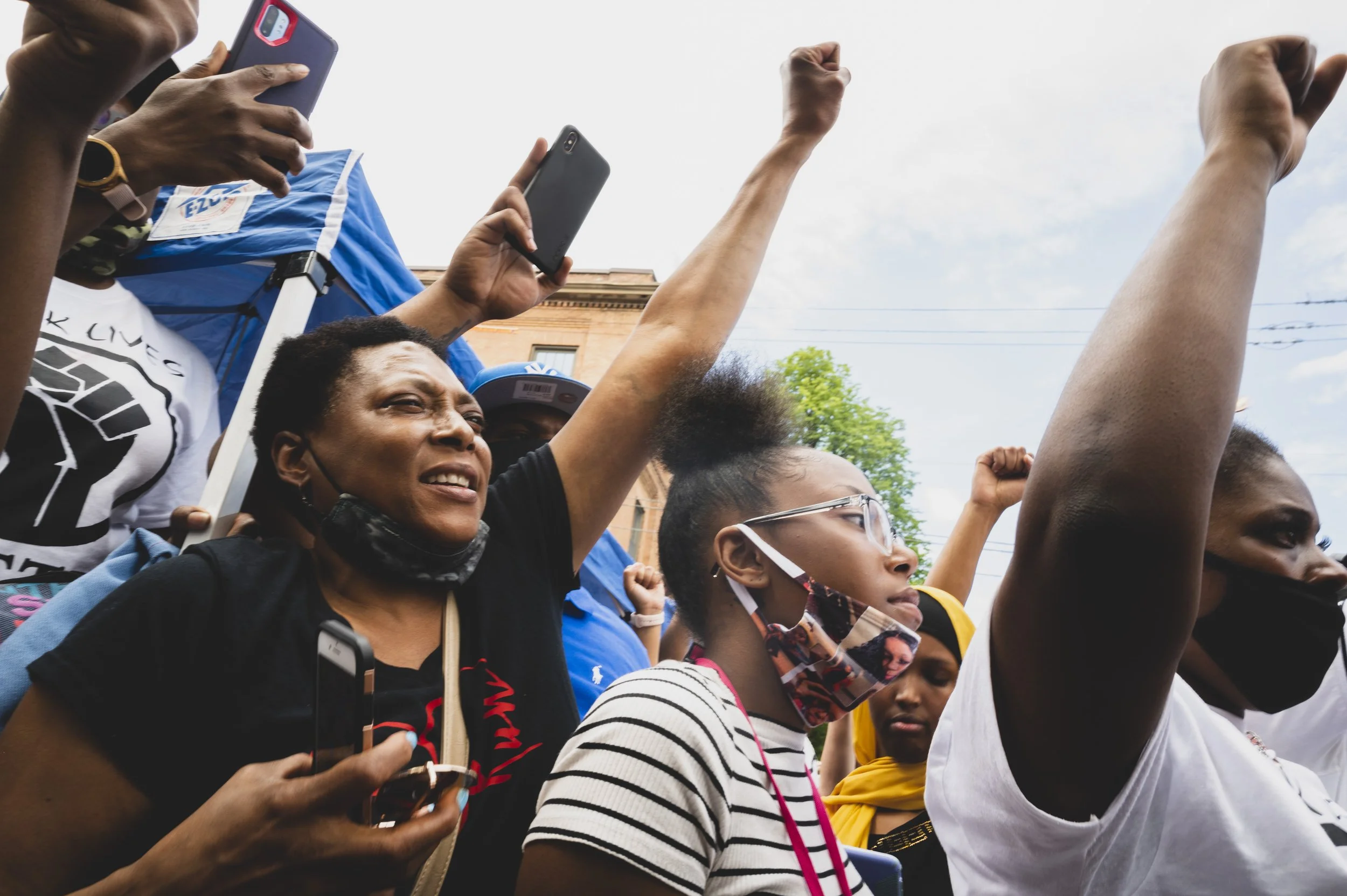 Group of protesters with raised fists, some wearing masks and holding phones, demonstrating during a rally.