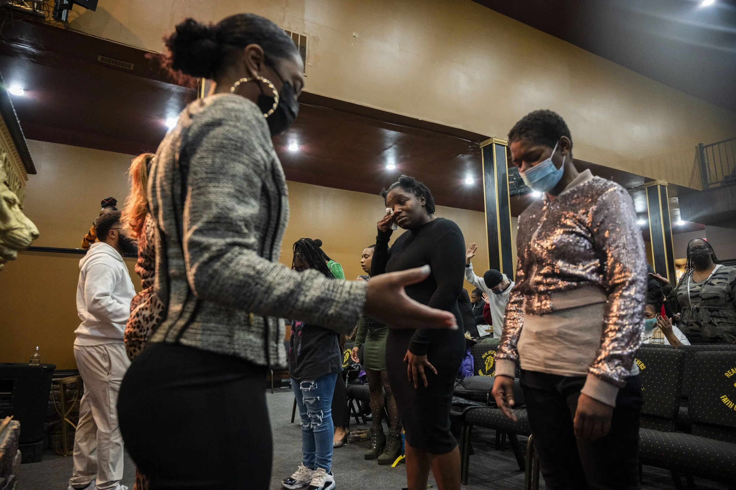 People standing in a circle with heads bowed and eyes closed, participating in a prayer or moment of reflection during a gathering in a dimly lit room.