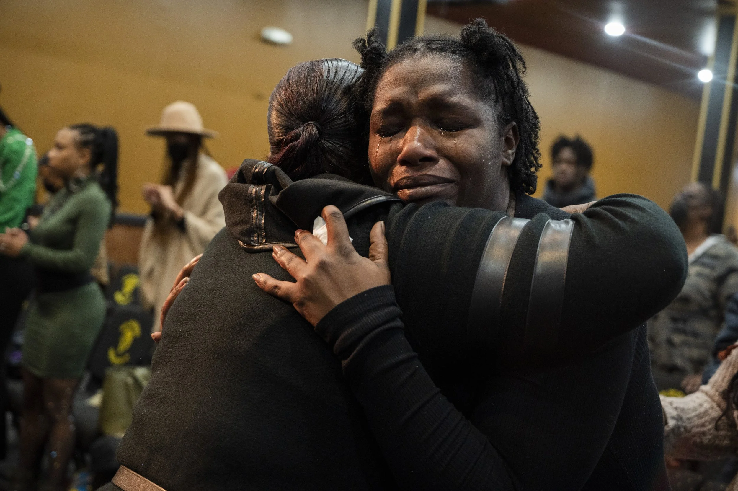 Two women hugging each other emotionally in a room filled with people, some with their eyes closed or looking down, during a heartfelt moment.