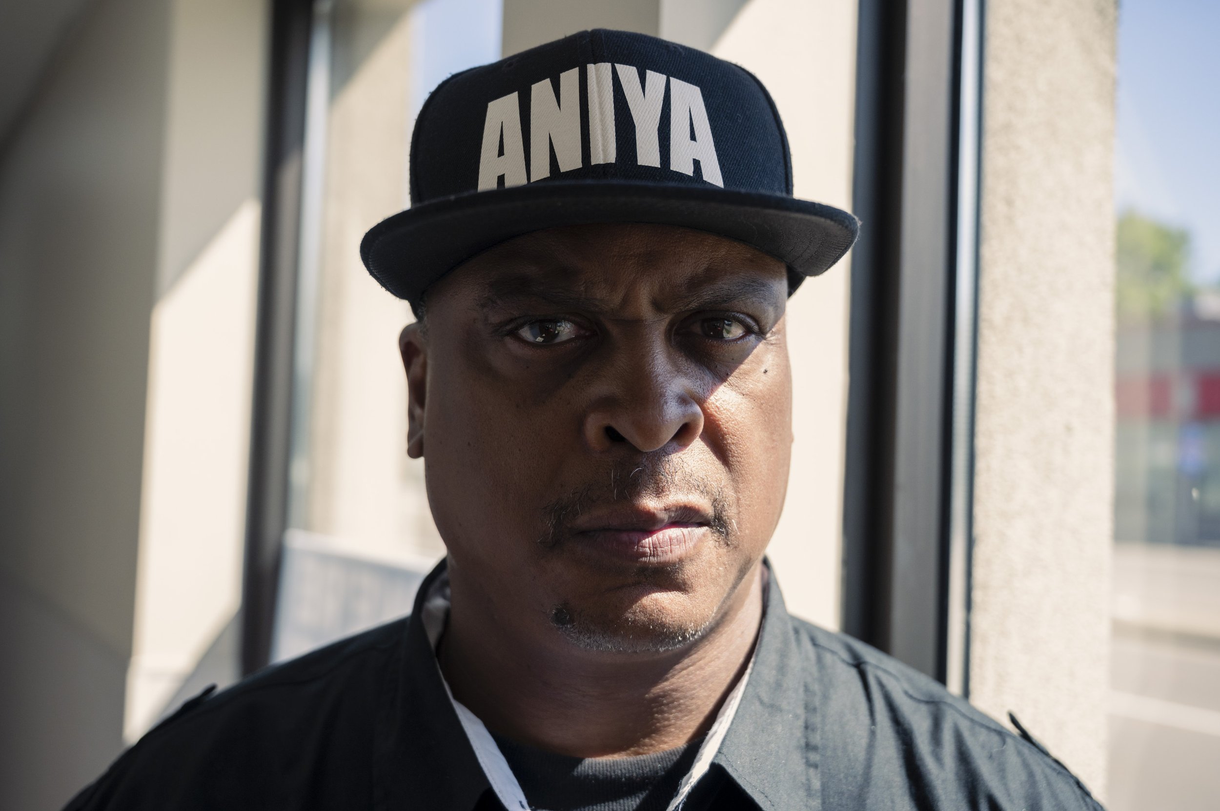 Close-up of a man with dark skin wearing a black cap with white text and a black jacket, standing inside near a window with sunlight in the background.