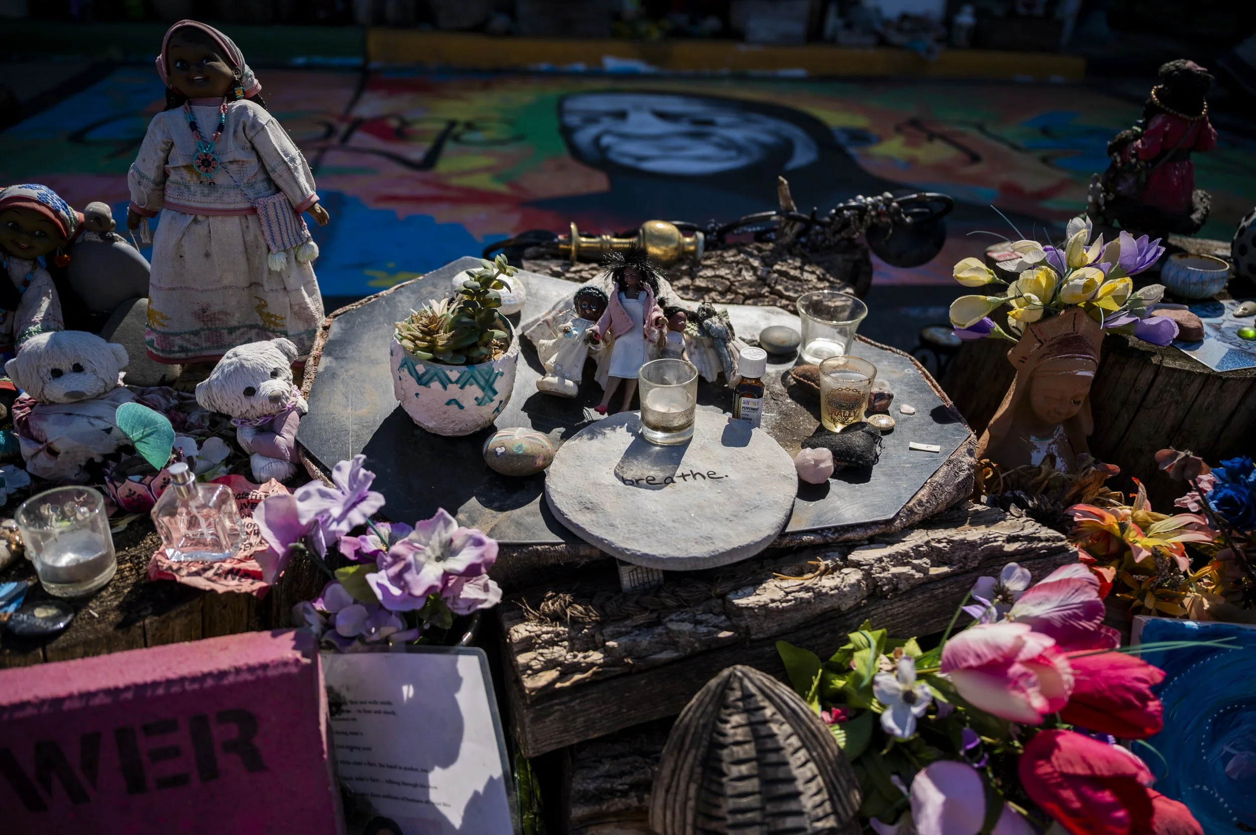 A memorial altar with handmade dolls, flowers, candles, and stones, with the word 'breathe' written on a stone, suggesting a tribute or remembrance.