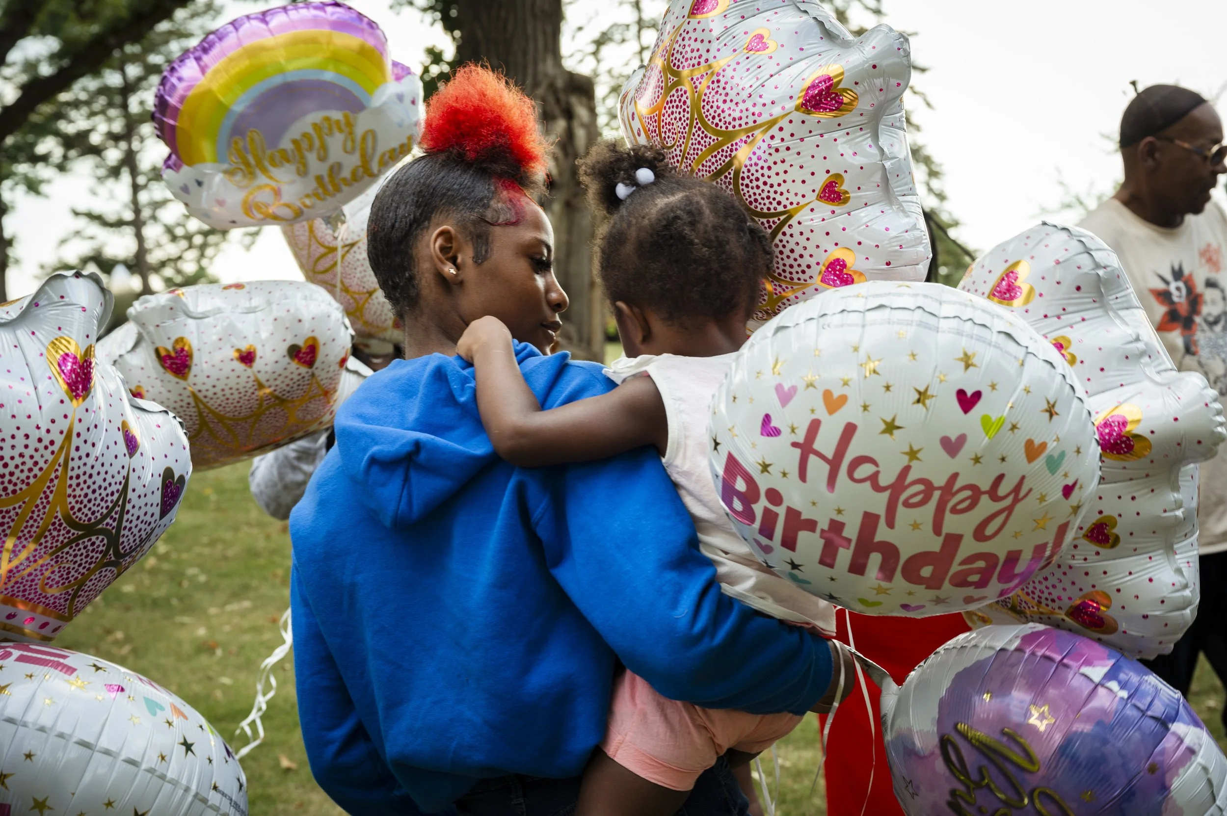 A Black teenager holding a young Black girl surrounded by colorful 'Happy Birthday' balloons at a cemetery to celebrate the life of Aniya Allen.