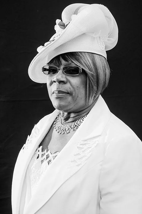 A woman wearing a wide-brimmed hat with decorative flowers, sunglasses, a necklace, and a white blazer against a dark background.