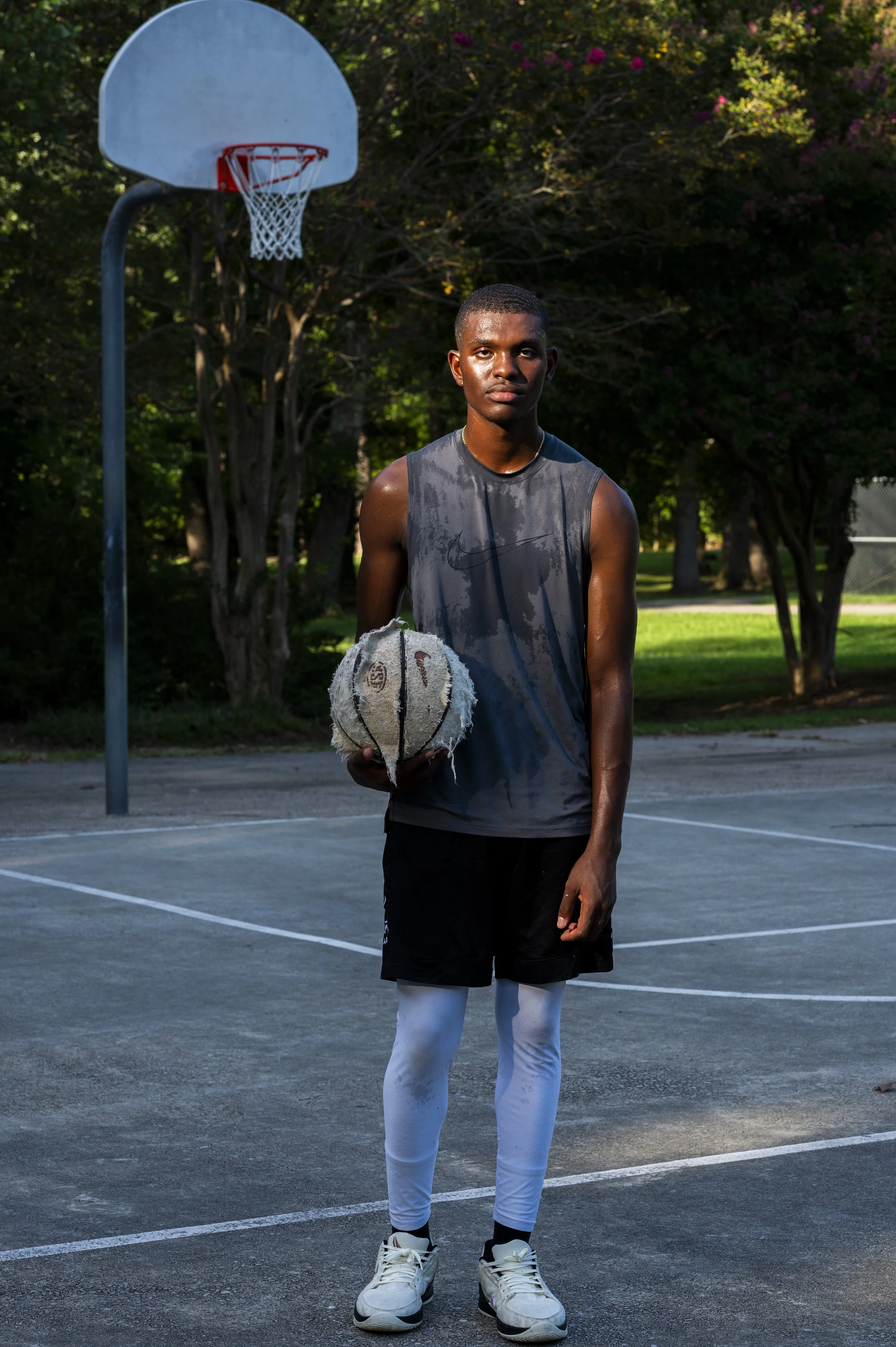 A young Black man standing on an outdoor basketball court at sunset, holding a worn basketball, wearing athletic clothes and looking at the camera. He is sun and sweat-drenched.