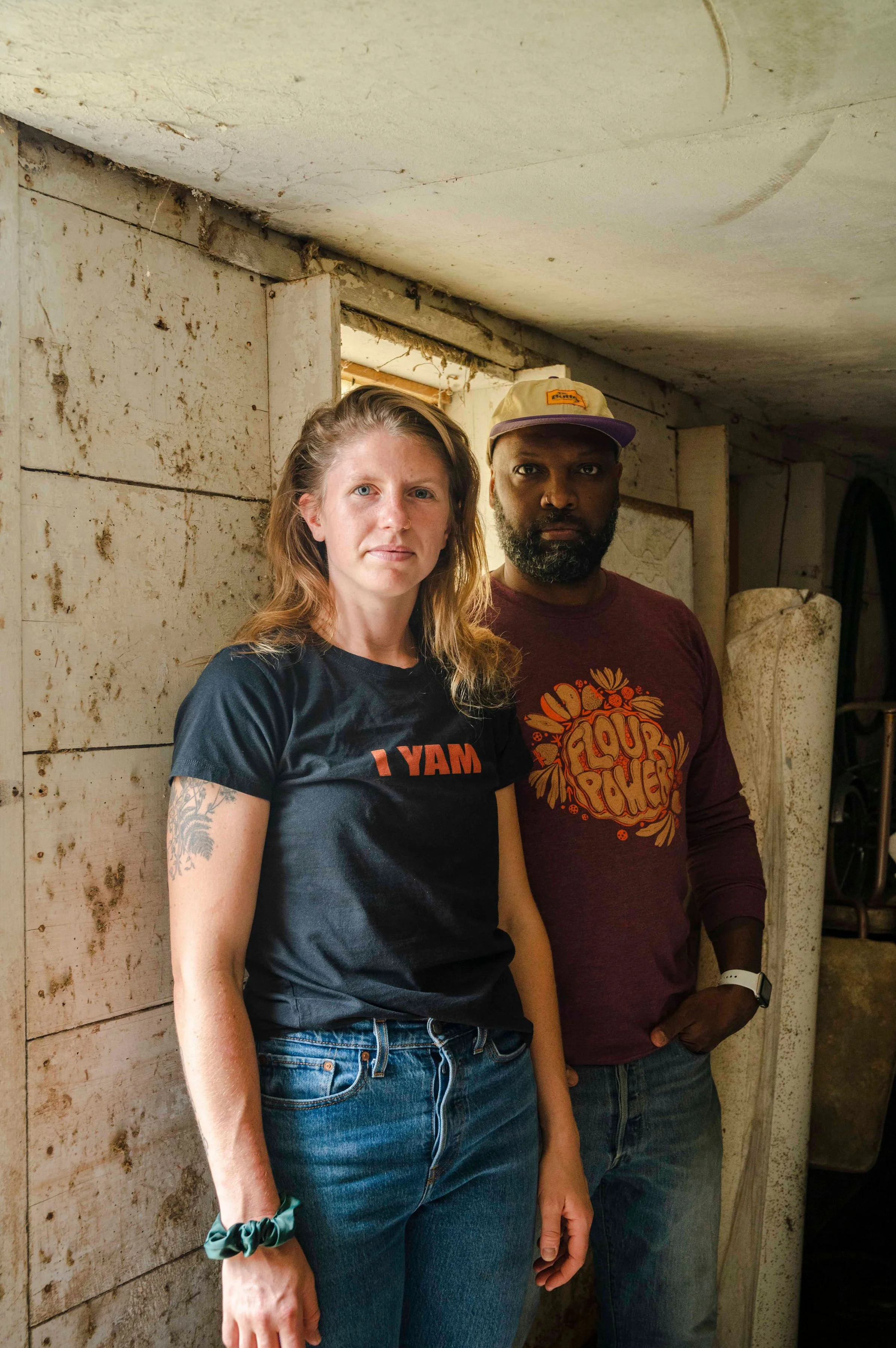 A woman with blonde hair and a tattoo on her left arm standing next to a man with a beard, both facing the camera. They are indoors in a rustic setting with weathered white wooden walls and a small window behind them. The woman is wearing a black t-s