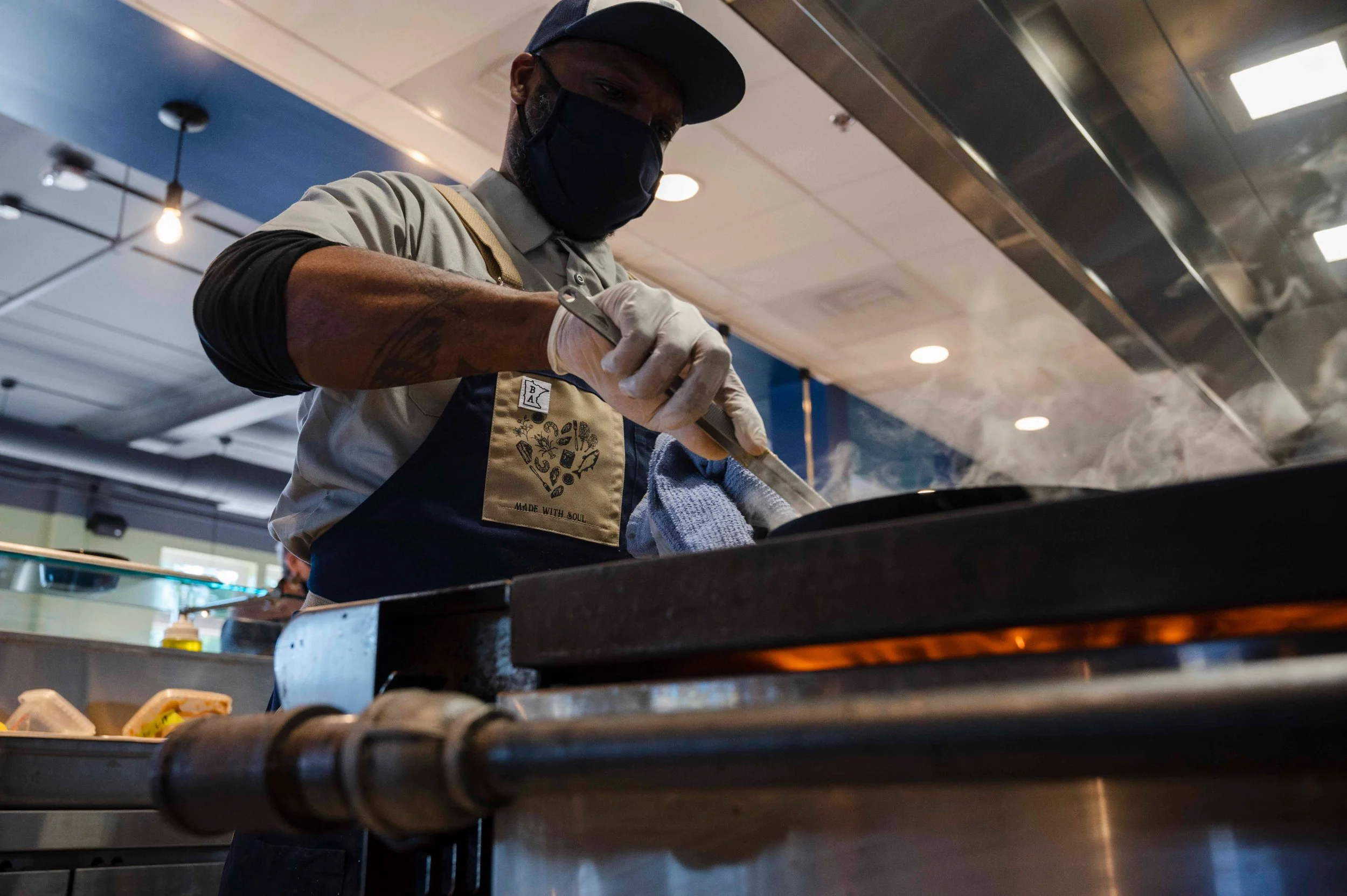 A man wearing a face mask and apron cooking on a griddle in a restaurant kitchen.