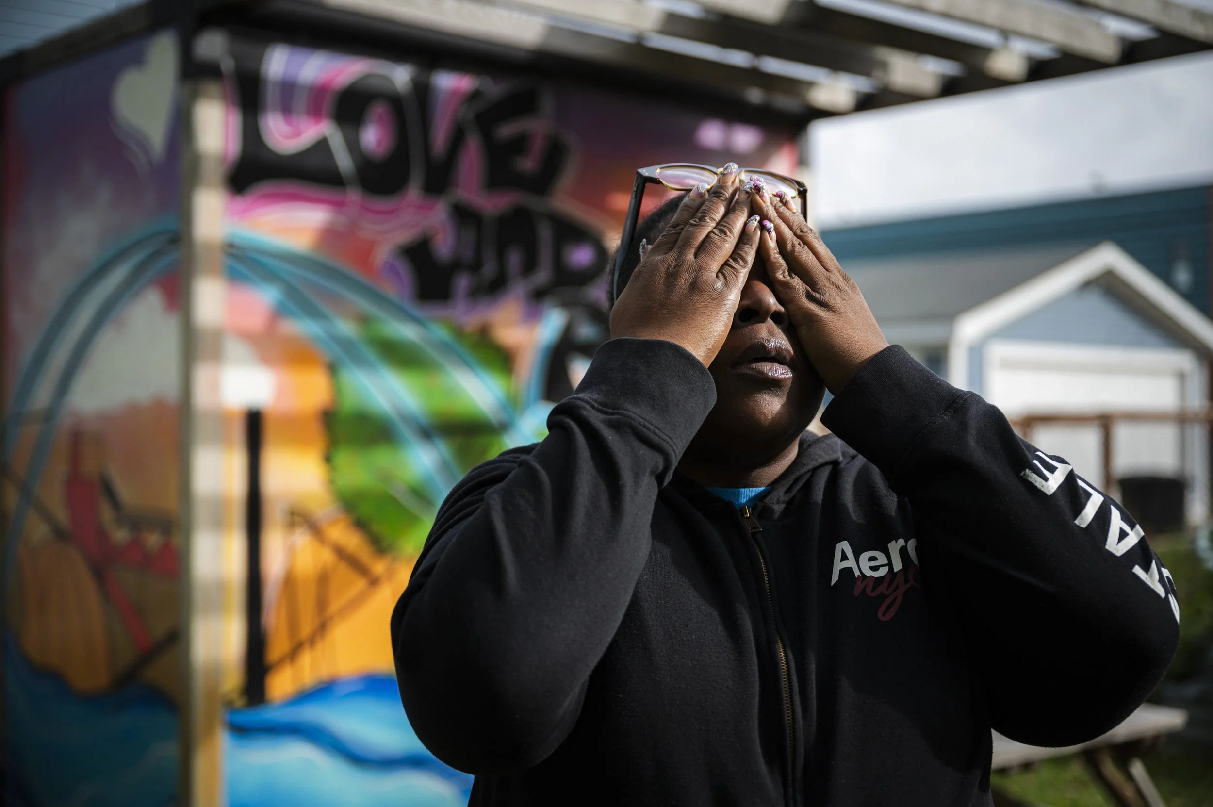 A woman with glasses is outdoors, covering her eyes with her hands, standing in front of a colorful graffiti wall.