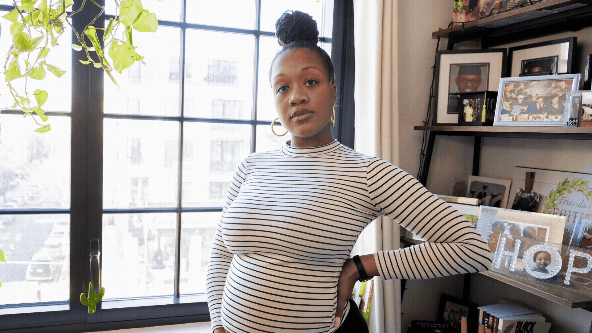 A woman with brown skin and hair styled in a high bun, wearing a white and black striped long-sleeve shirt, standing in a room with large window and shelves filled with framed photos and decorative items.