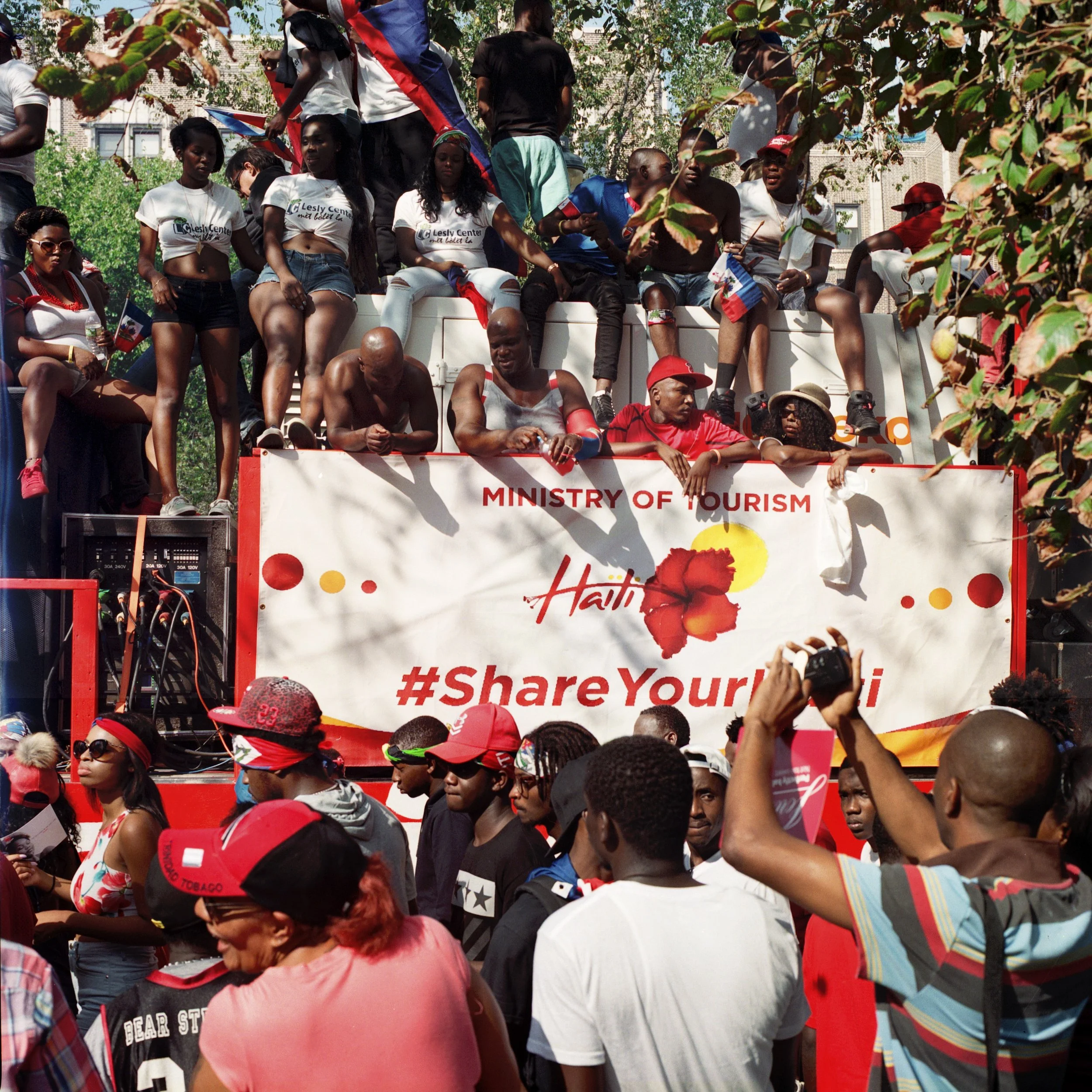 A crowd of people participating in a parade or festival in Haiti, with some standing on a float that has a banner reading 'Ministry of Tourism Haiti #ShareYourHaiti'. The float is decorated with a large Haitian flag and colorful flowers, with people 