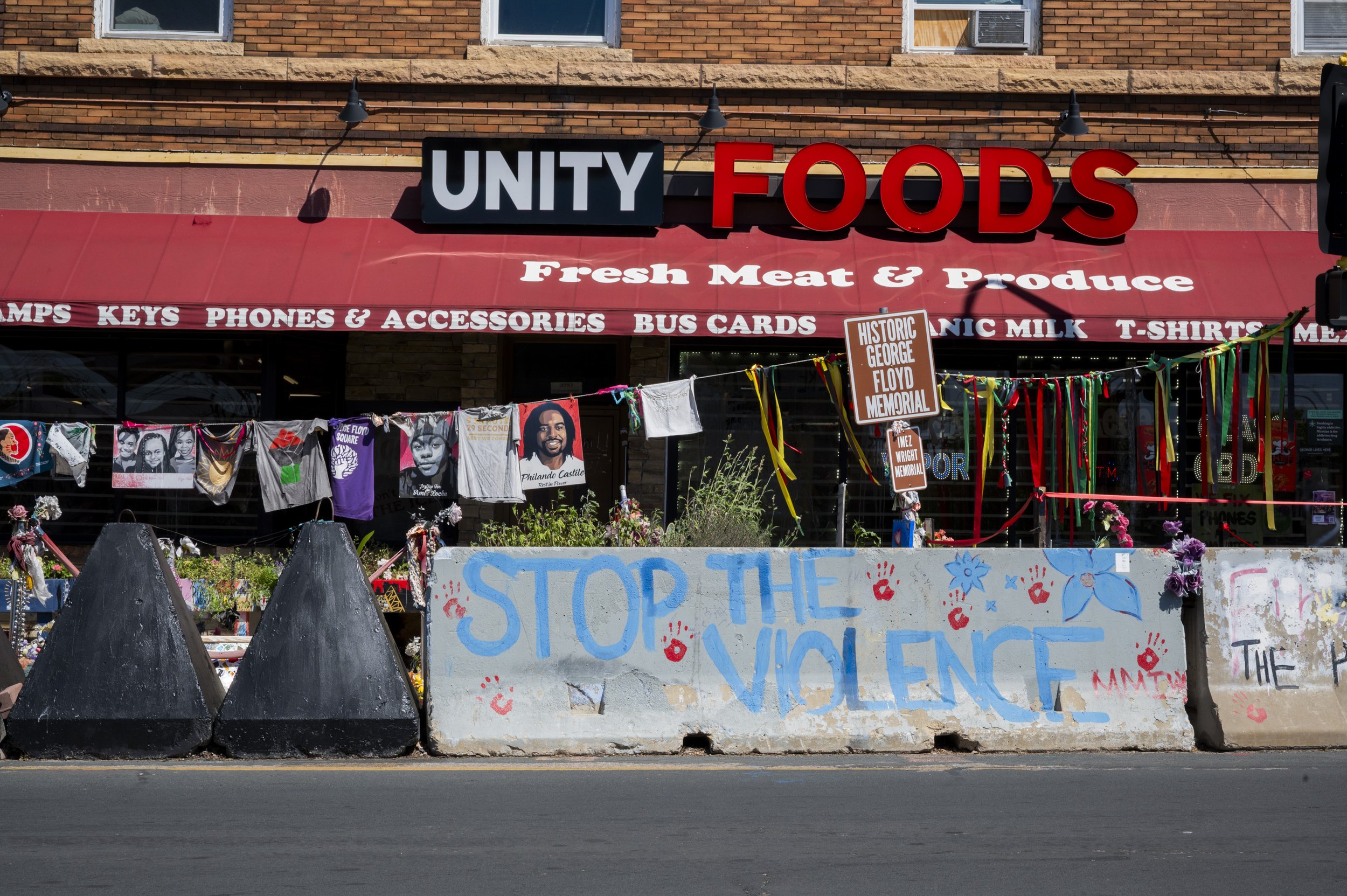 Graffiti on a concrete barrier reading 'STOP THE VIOLENCE' with red handprints, in front of a storefront labeled 'UNITY FOODS' selling fresh meat and produce, with memorial signs, posters, colorful ribbons, and hanging laundry.