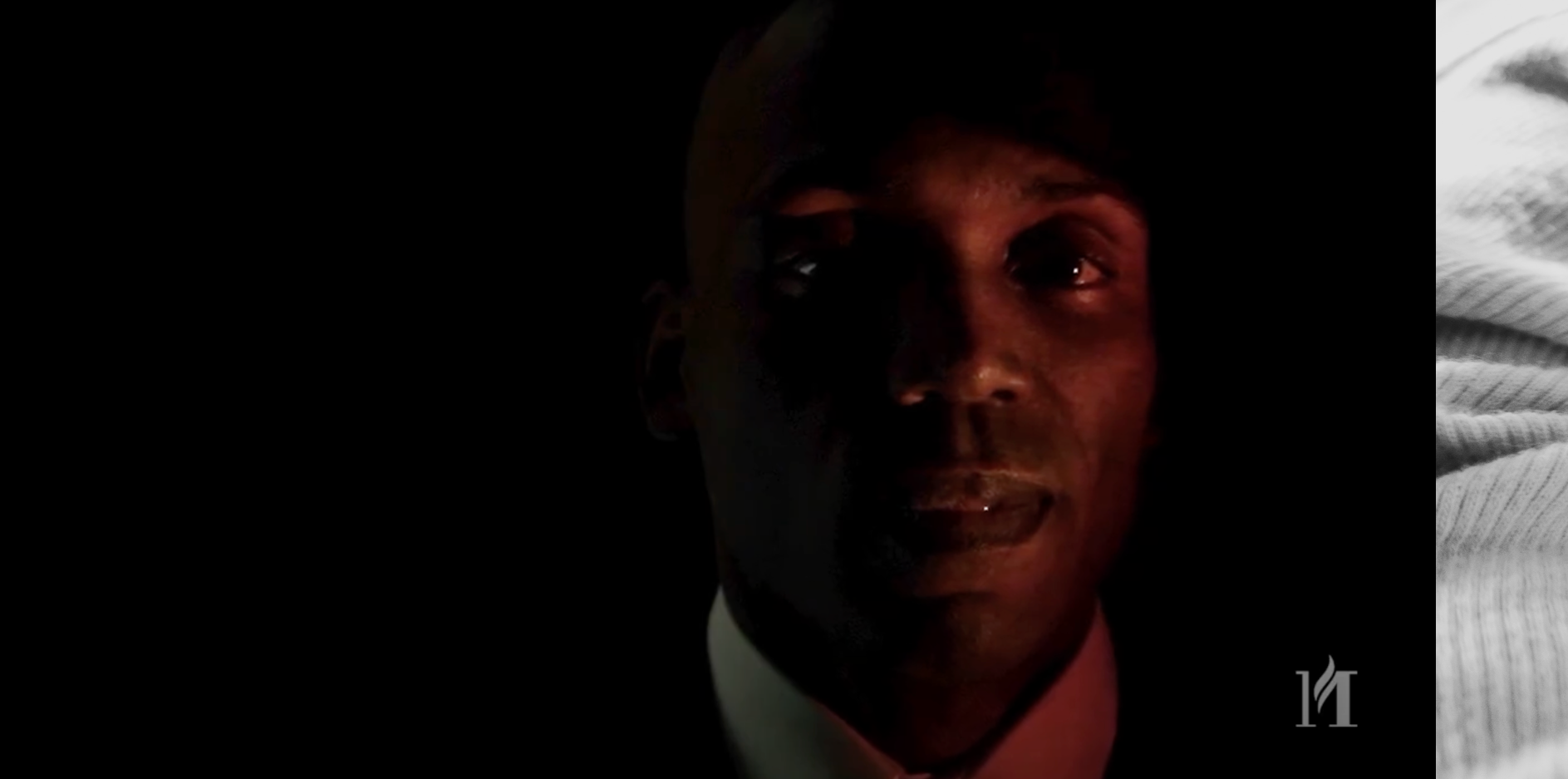 A portrait of a man with dark skin, smiling slightly, wearing a light-colored shirt and dark jacket. The background is black, and there is a faint watermark in the lower right corner.