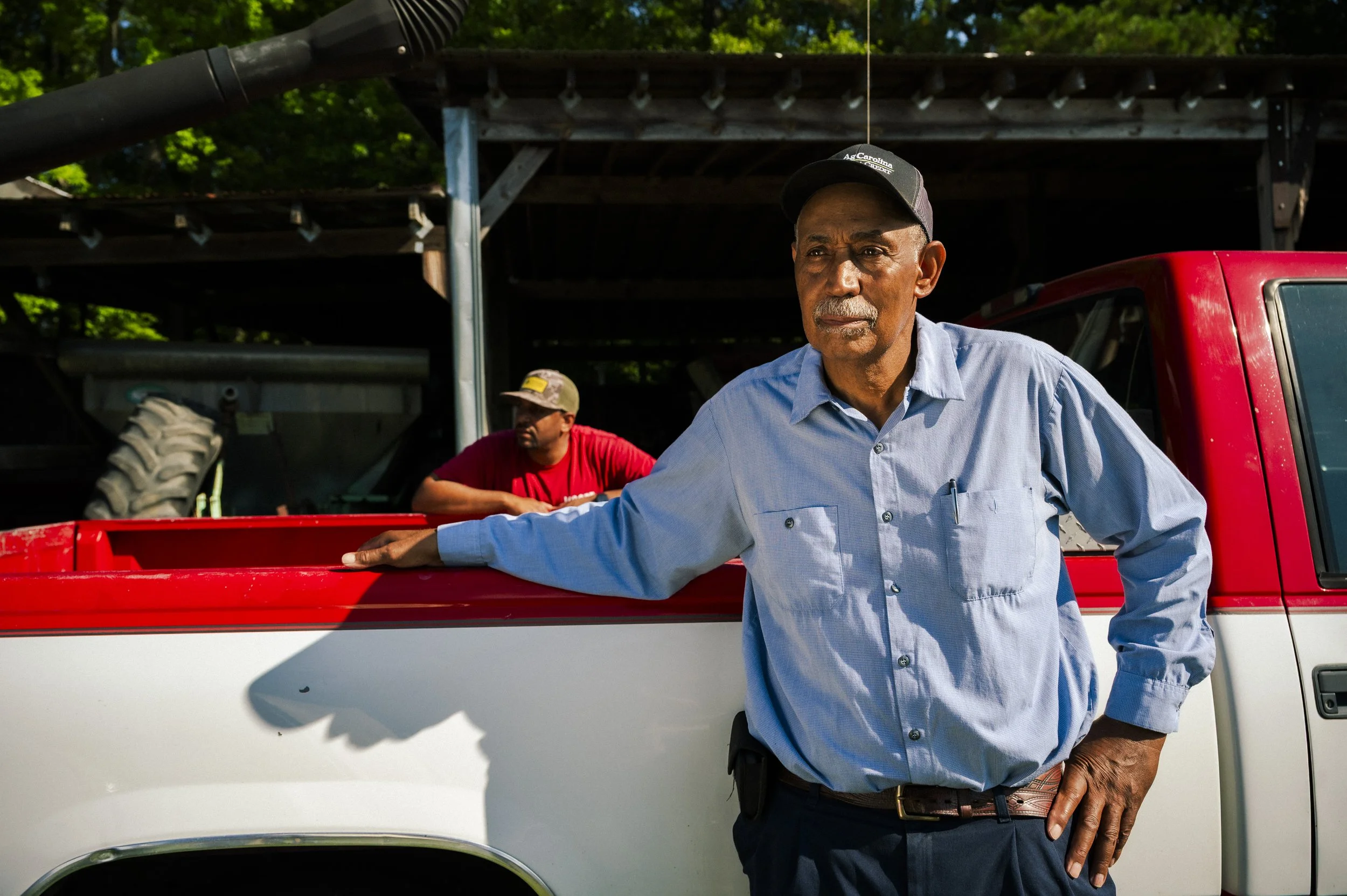 An older man with a gray mustache and serious expression stands leaning on the side of a red pickup truck. He's wearing a light blue button-up shirt, a black belt, and a black cap. In the background, there is another man wearing a red shirt and a bei
