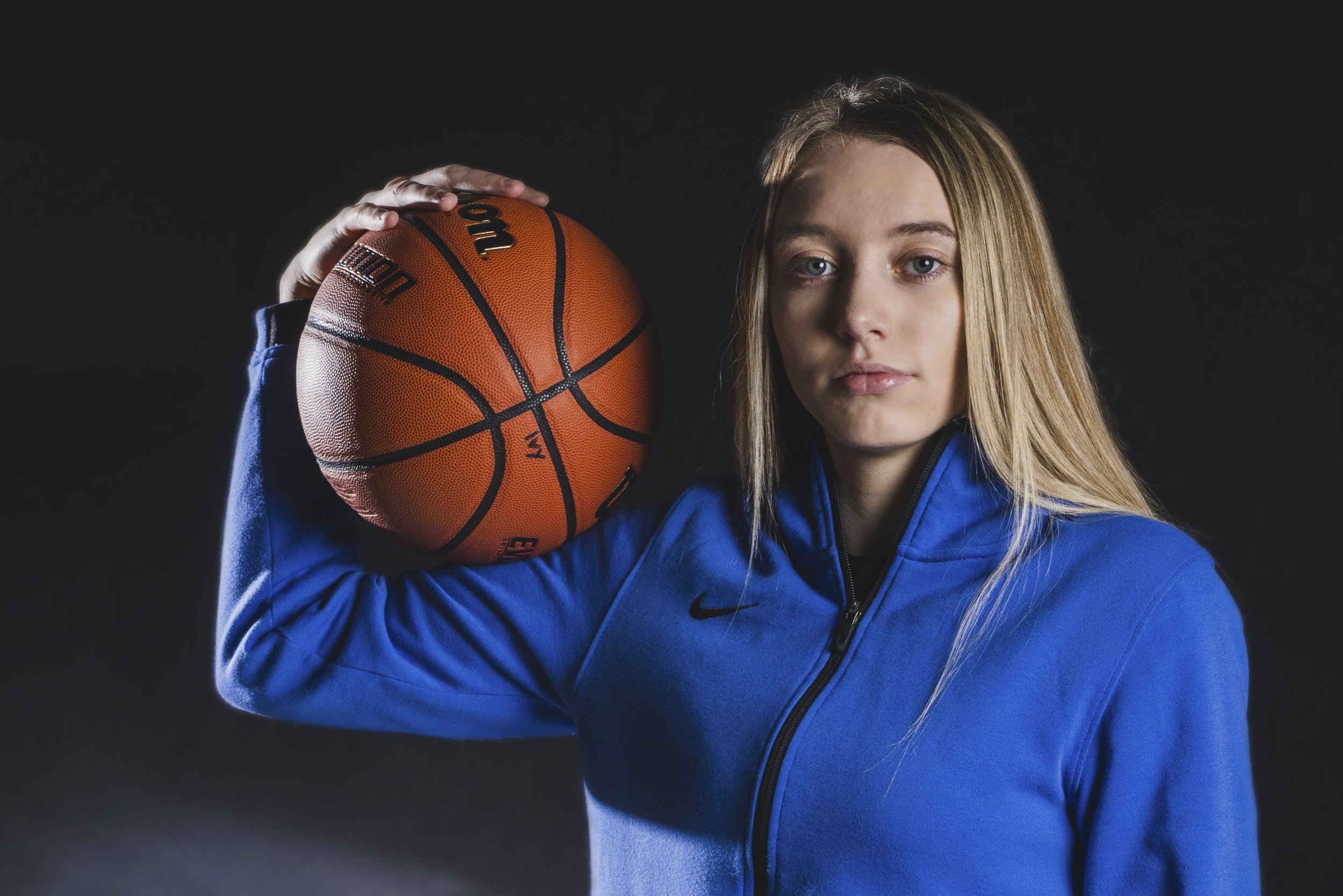 Young woman with long blonde hair holding a basketball over her shoulder, wearing a blue sports jacket, against a dark background.
