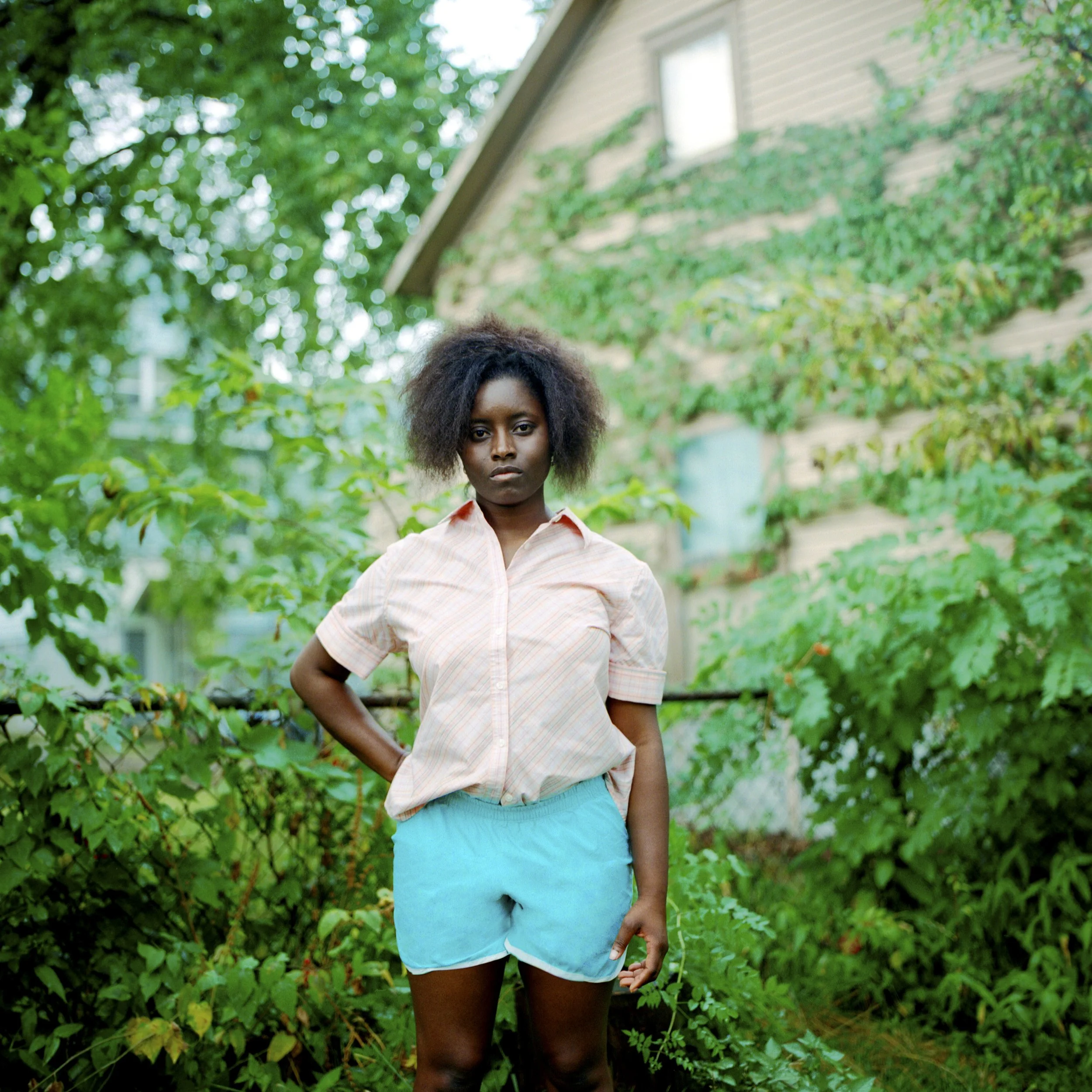 A young woman standing outdoors in front of lush greenery and a house covered in vines, looking serious at the camera, wearing a light pink shirt and blue shorts.
