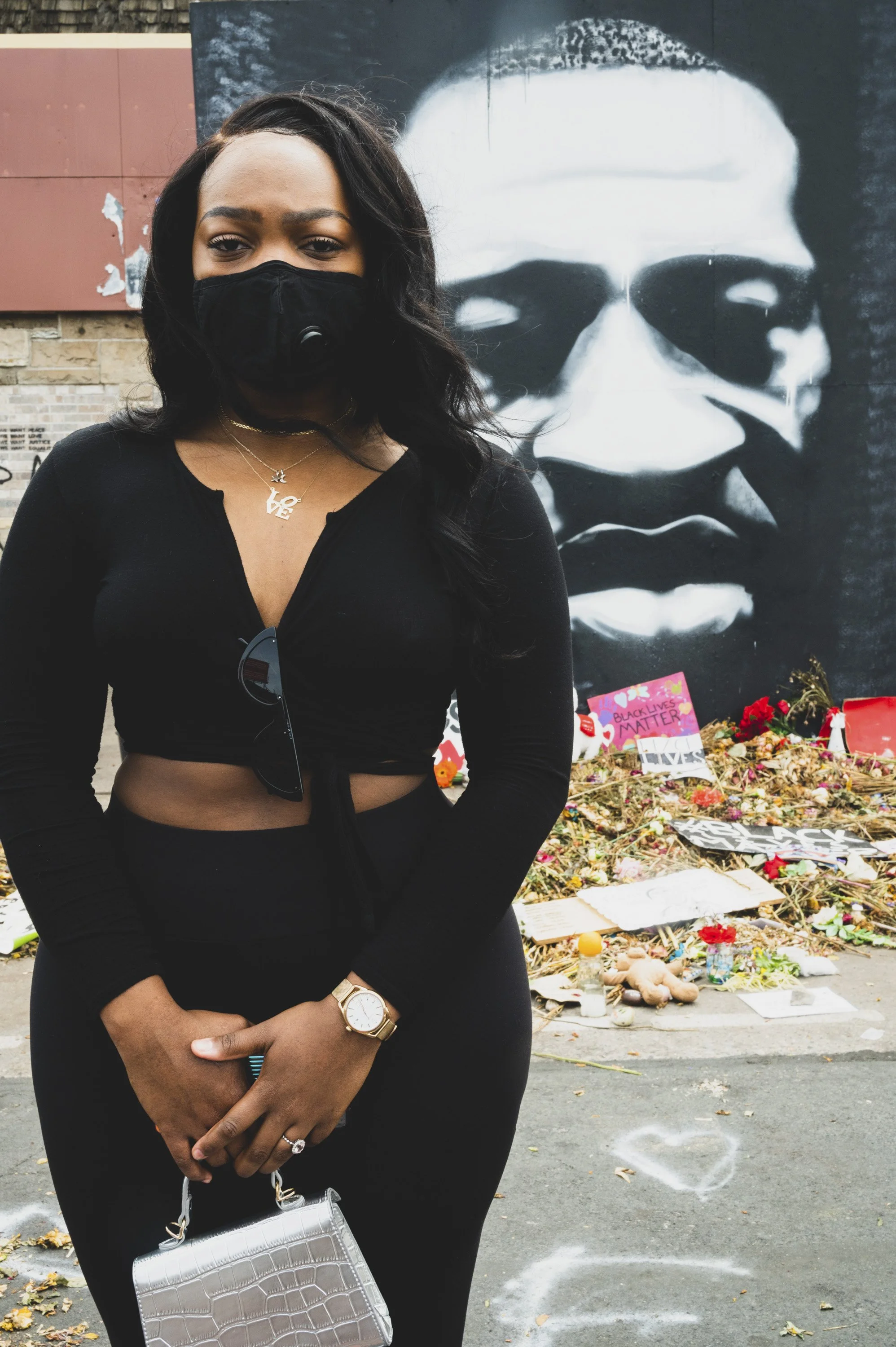 A woman standing in front of a mural of a face with sunglasses and a mask. She is wearing a black long-sleeve top, a gold watch, layered necklaces that say 'LOVE' and a star, and is holding a silver handbag. There are memorial items and flowers on th