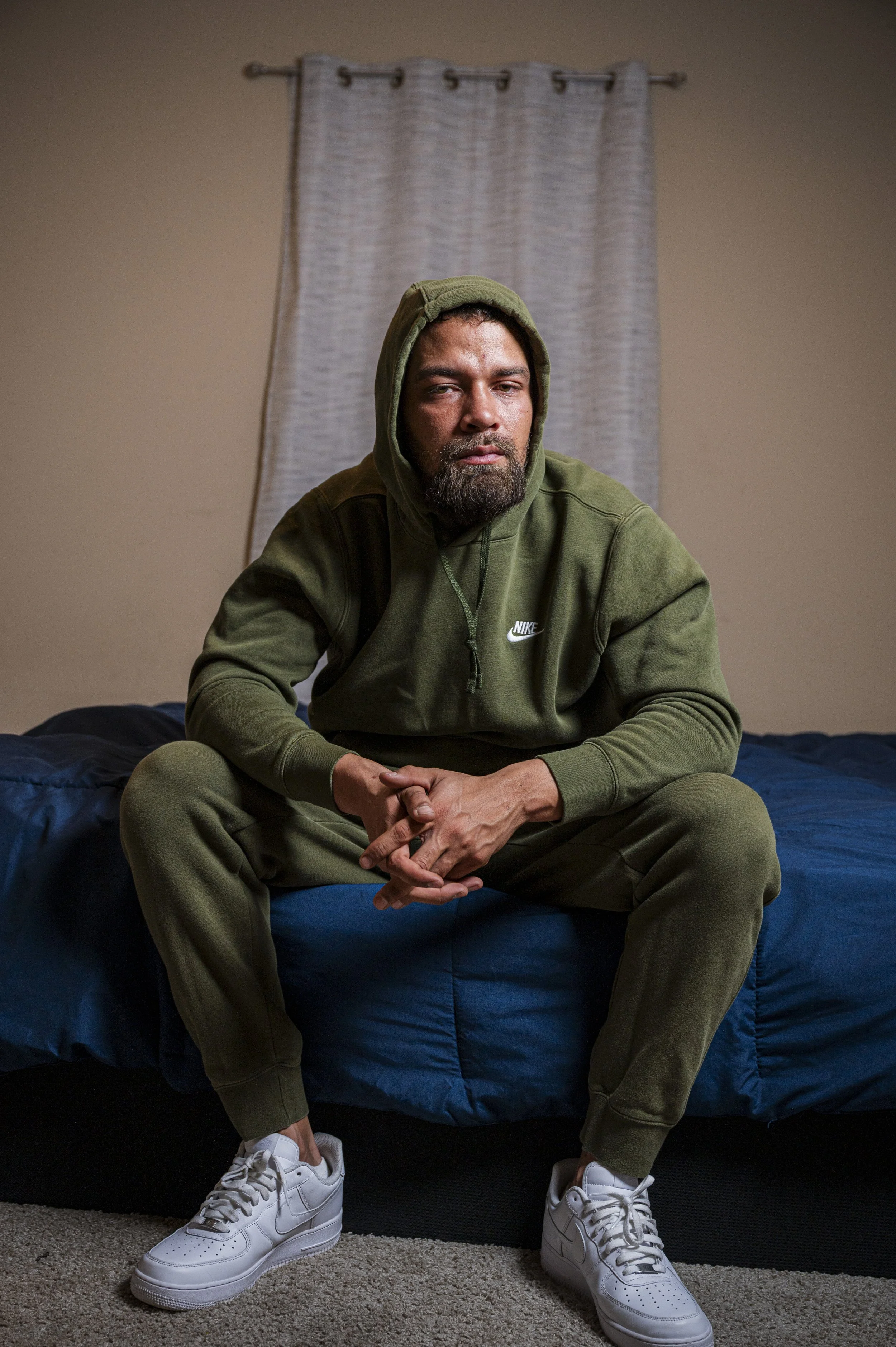 A man in a green hoodie and sweatpants sitting on a bed in a room with beige walls, a blue blanket, and a window with waves.