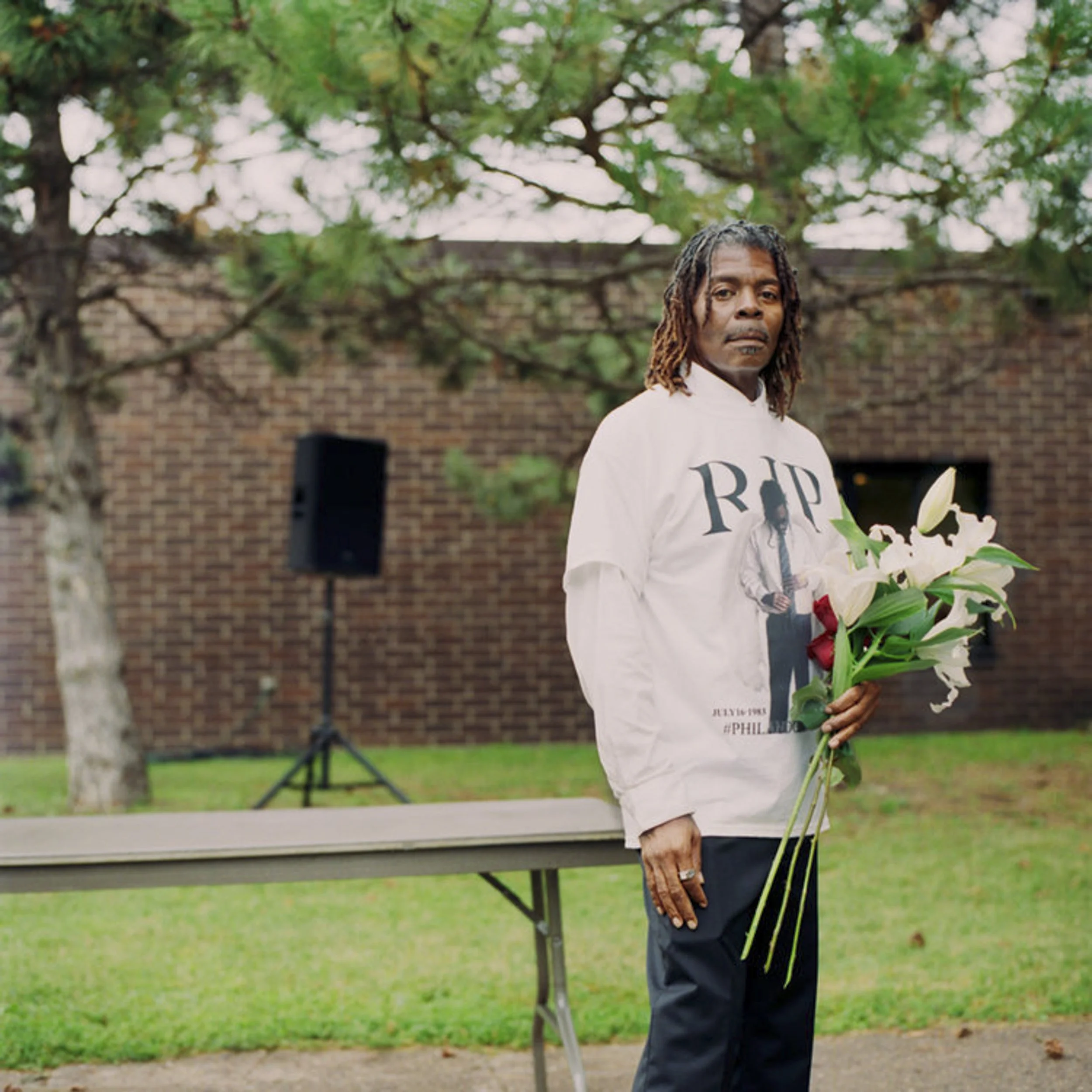 A man with dreadlocks wearing a white shirt with 'RIP' on it standing outdoors near a floral arrangement, with a brick wall and green trees in the background.