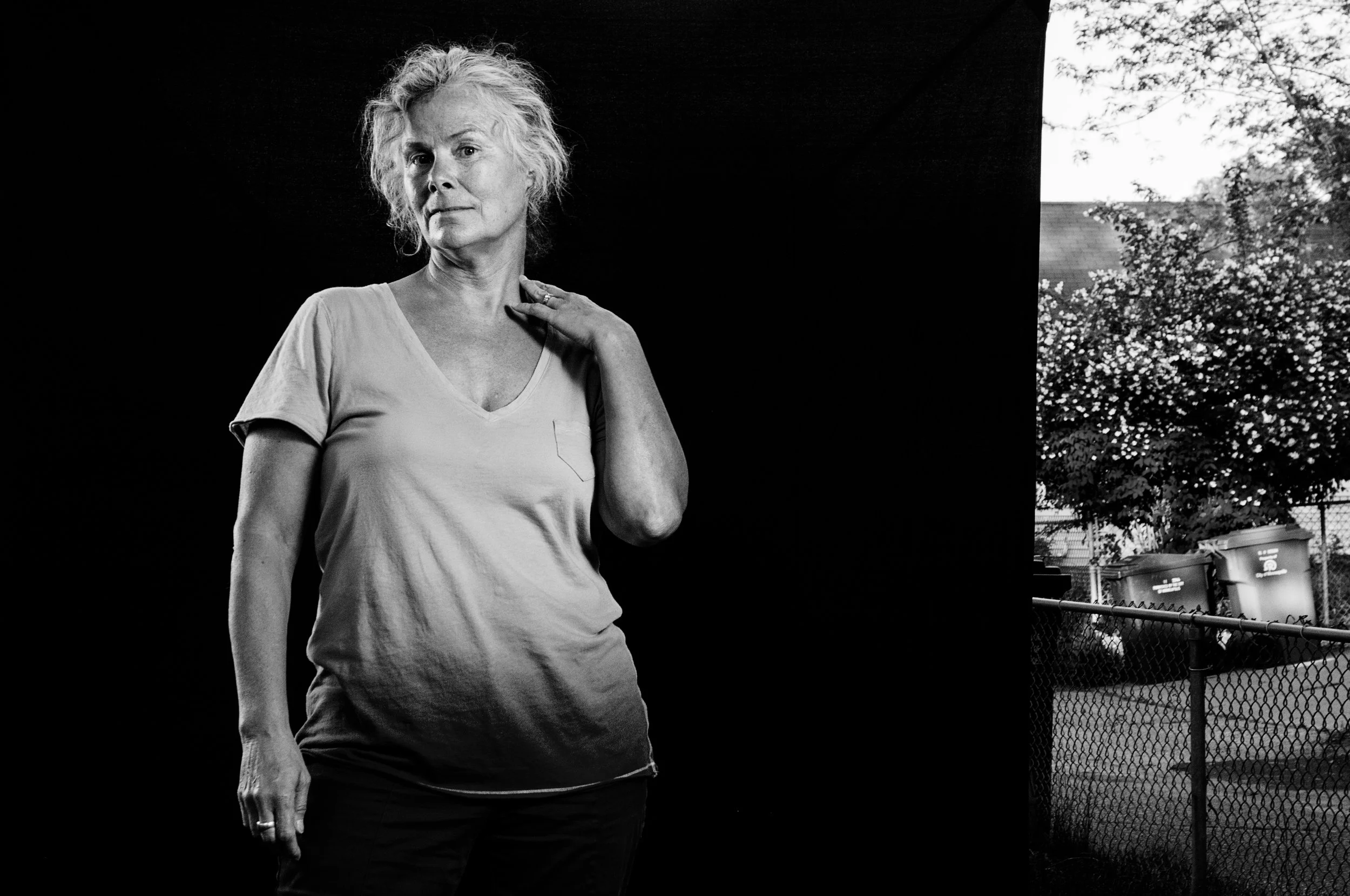 A black and white photo of an older woman with curly hair, wearing a t-shirt, standing against a dark background with a portion of a chain-link fence and trees visible in the background.