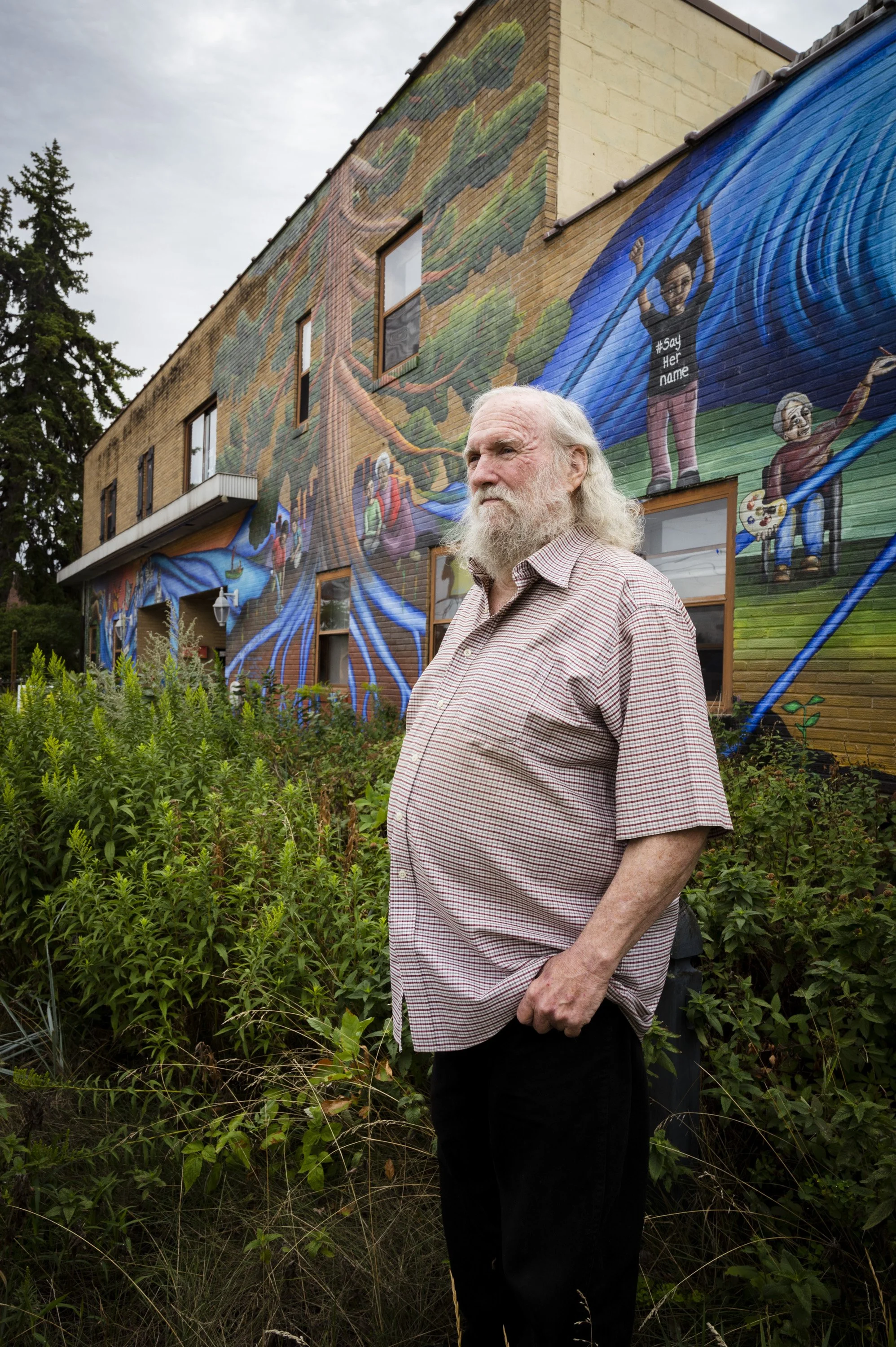 An elderly man with long gray hair and a beard standing amidst green bushes in front of a building with a colorful mural depicting trees and children playing. The man is wearing a short-sleeved checkered shirt and black pants, looking thoughtfully to
