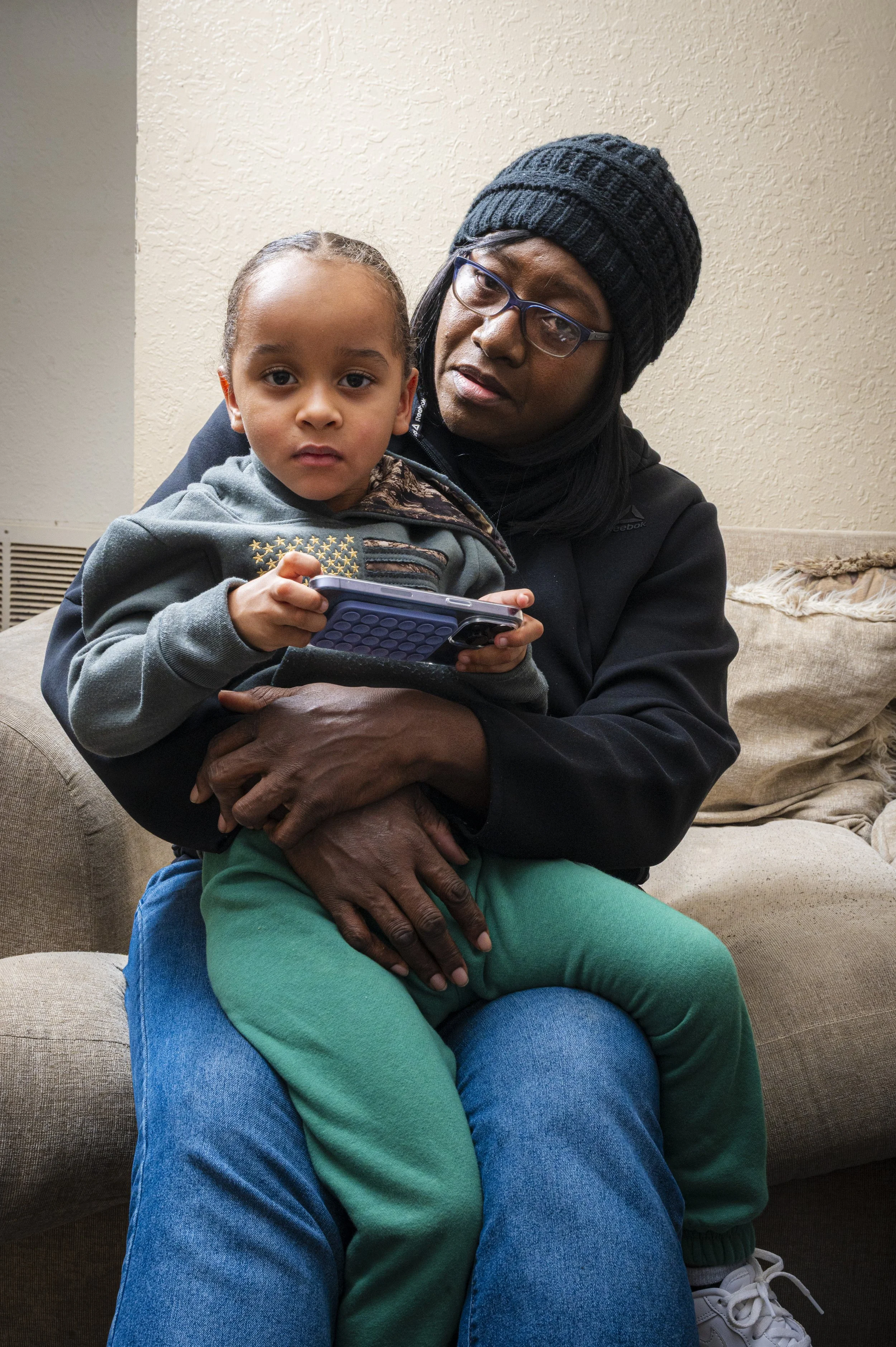 A woman, wearing glasses and a black beanie, sitting on a beige couch, holding a young girl on her lap. The girl is holding a smartphone and looking at the camera, while the woman looks at her with a concerned or attentive expression. The background 