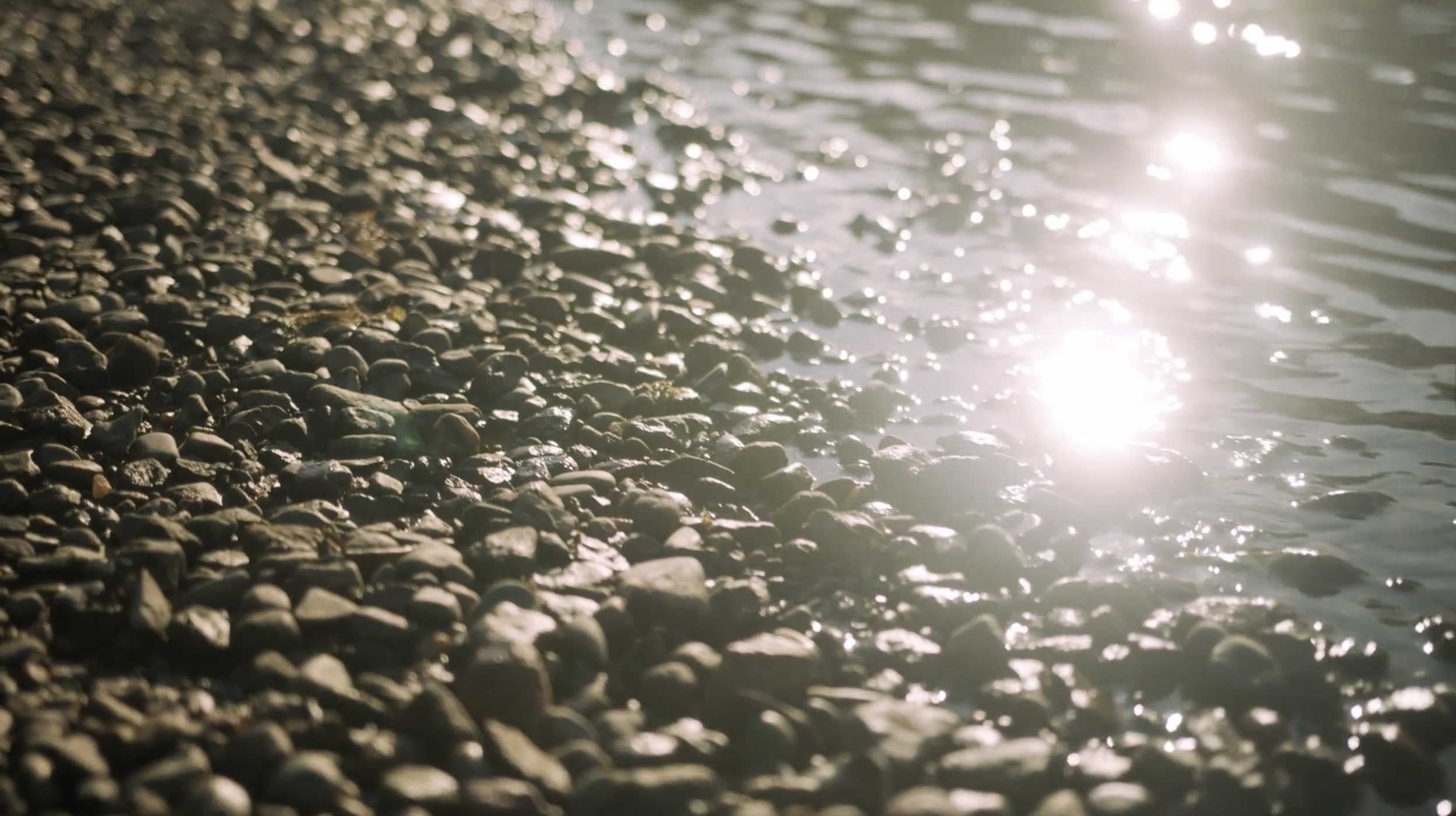 Sunlight reflecting off the surface of a pebble beach beside a body of water, with small rocks and stones along the shoreline. This is the base of the Mississippi River and the confluence of B'Dote.