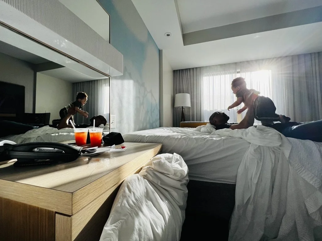 Two children playing on a bed in a hotel room with sunlight coming through the window. Drinks and a telephone are on the bedside table.