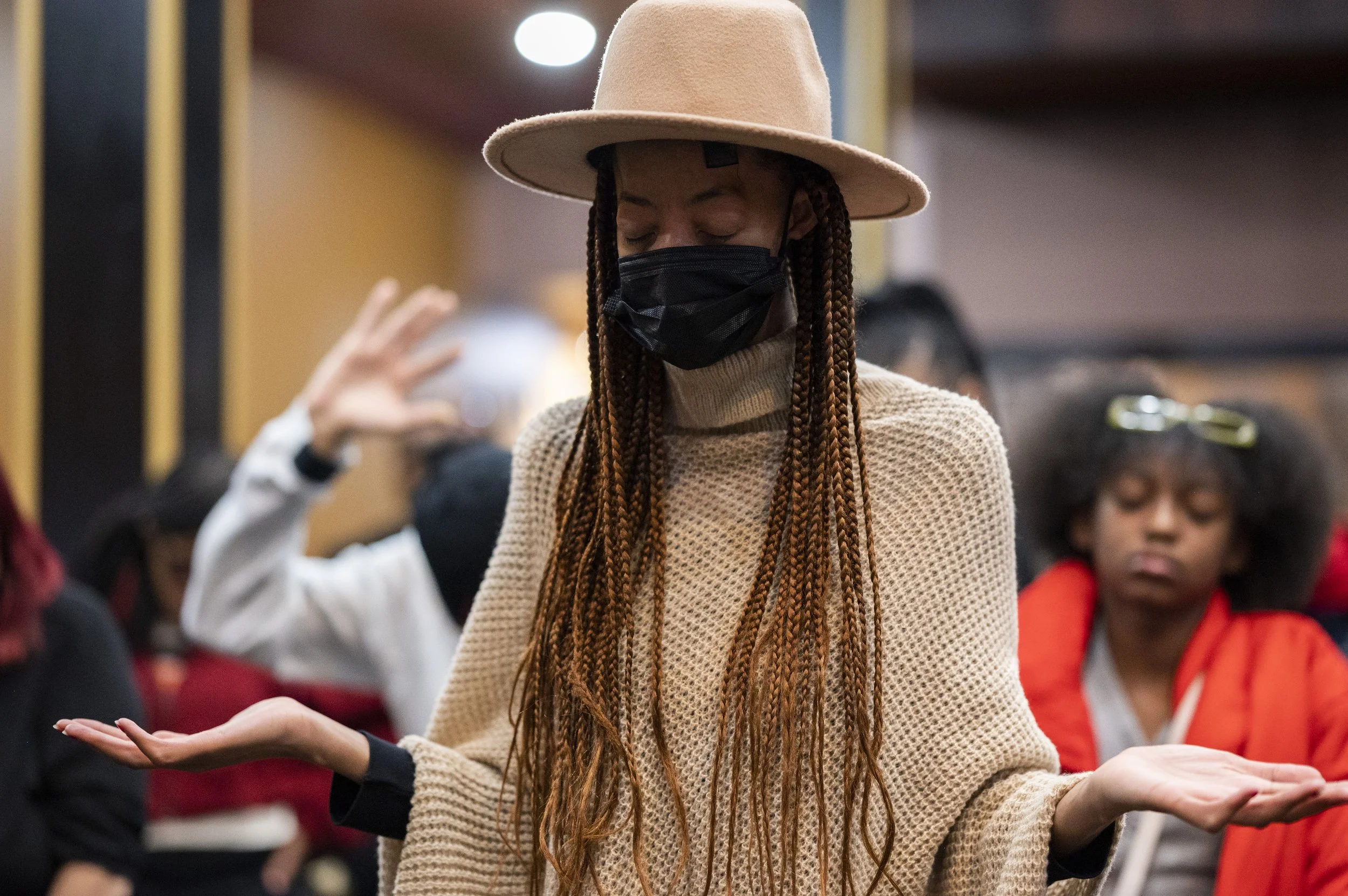 A woman with long braided hair wearing a beige sweater, a light pink wide-brimmed hat, and a black face mask, with her eyes closed and hands raised in a meditation or prayer position, standing among other people in a group setting.