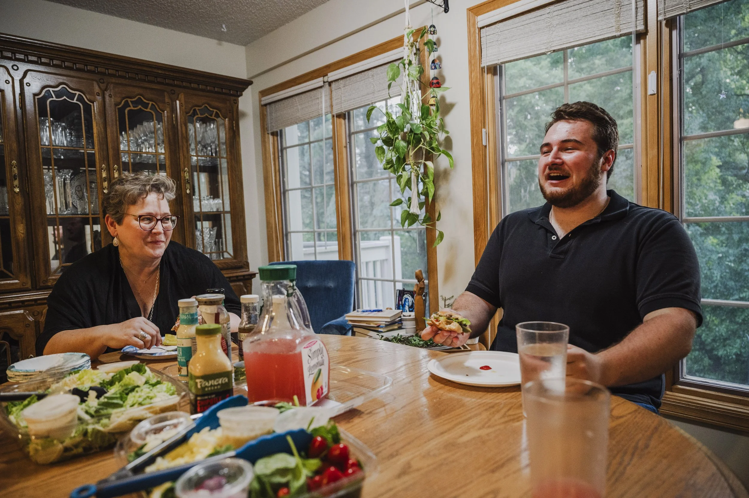 A man and a woman sitting at a dining table during a meal. The woman has short gray hair and glasses, and the man has short dark hair and a beard. There are various food items, condiments, and drinks on the table. Large windows in the background reve