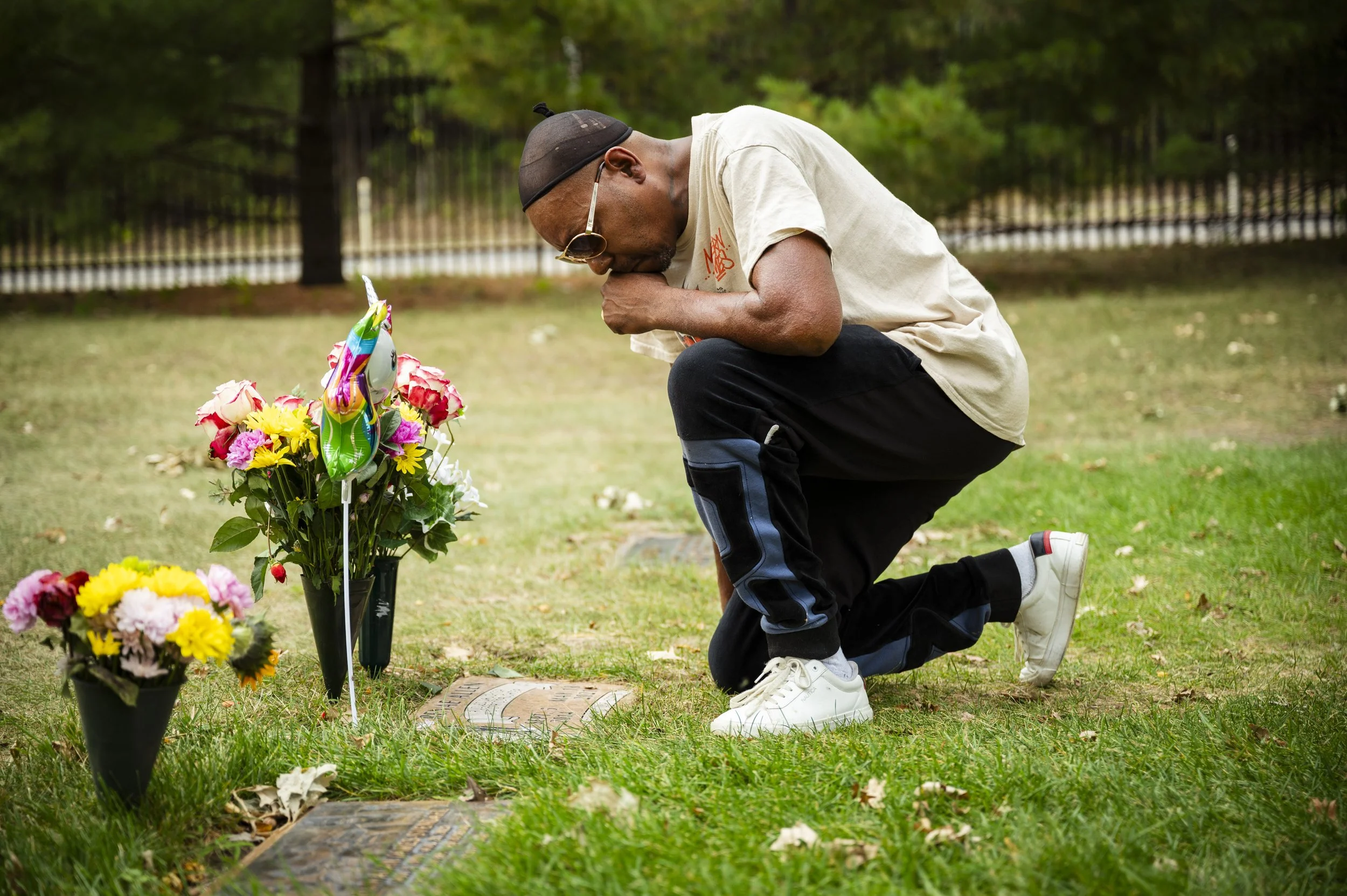 A man kneeling at a gravesite, praying with his head bowed and hand covering his mouth, next to flower arrangements.