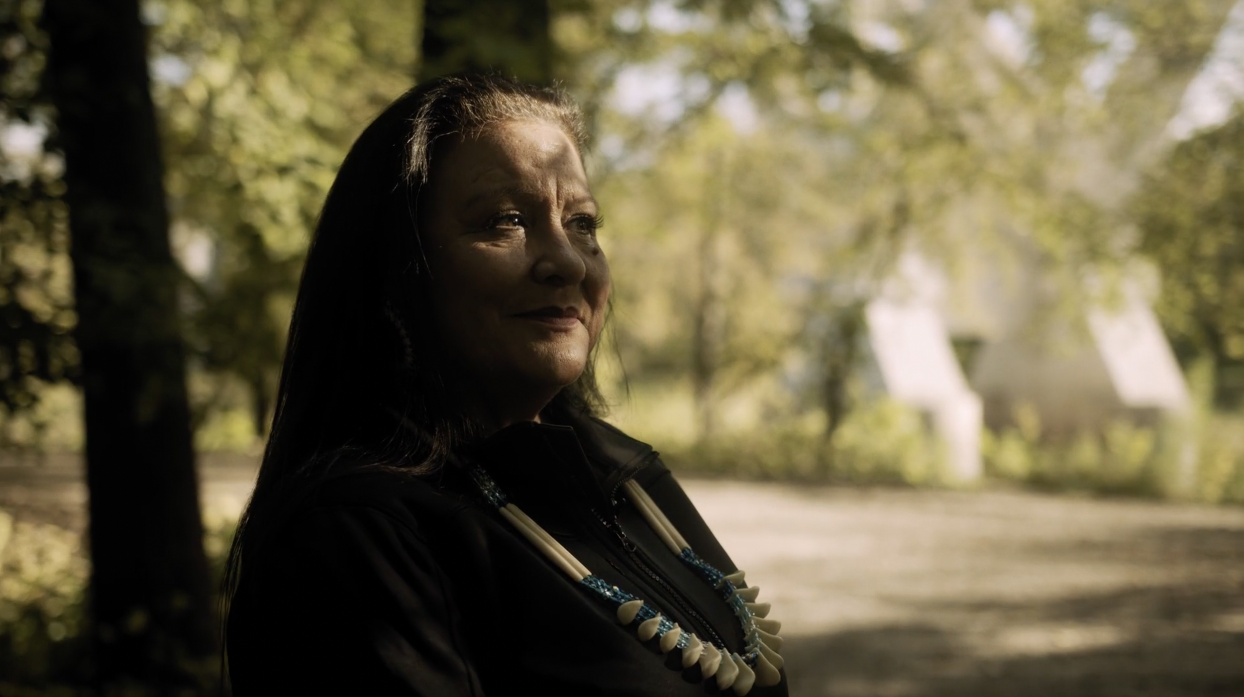 A Native American woman with long dark hair smiling outdoors during daytime, wearing a black jacket and a necklace made of shells and beads, with trees and white tents in the background.