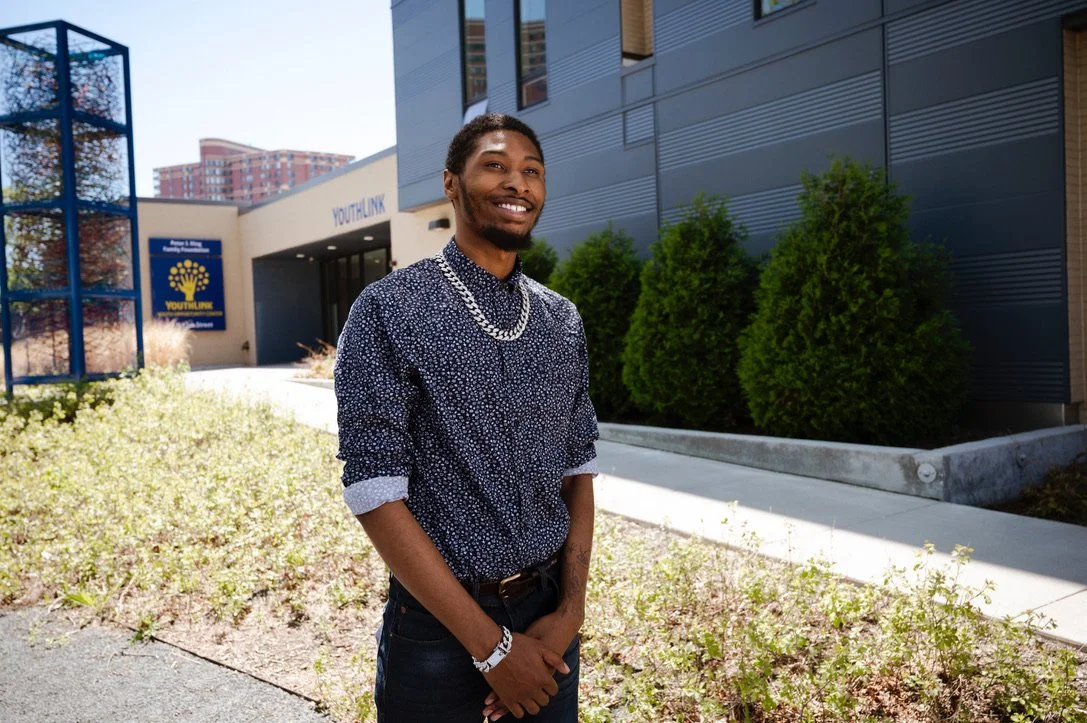 A smiling young Black man standing outside the YouthLink building with bushes and trees in the background.