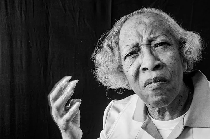 An elderly Black woman with curly gray hair appears confused or upset, wearing a light-colored shirt against a dark background.