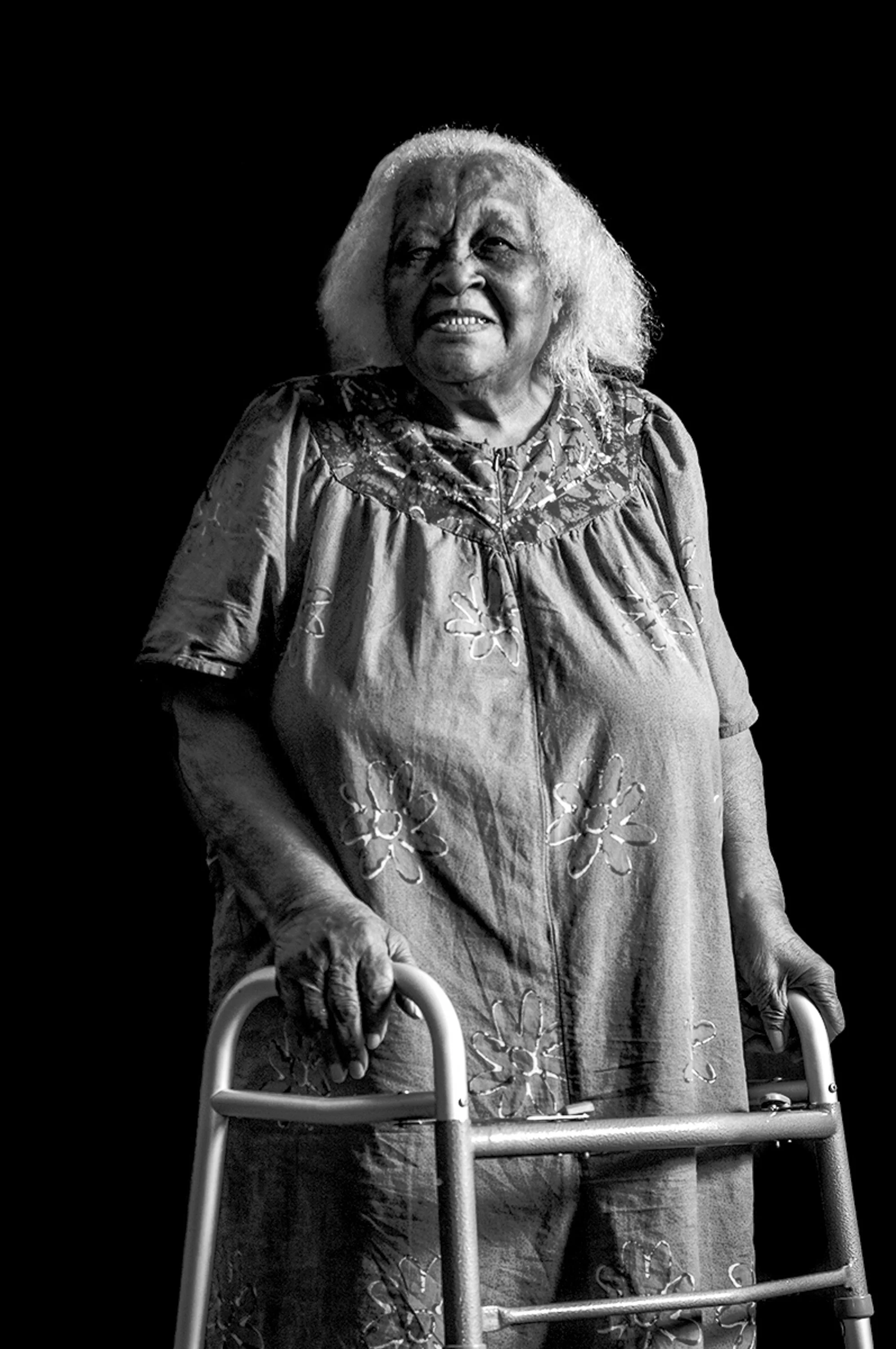 An elderly woman with curly gray hair smiling, holding a walker, wearing a floral dress, standing against a dark background.