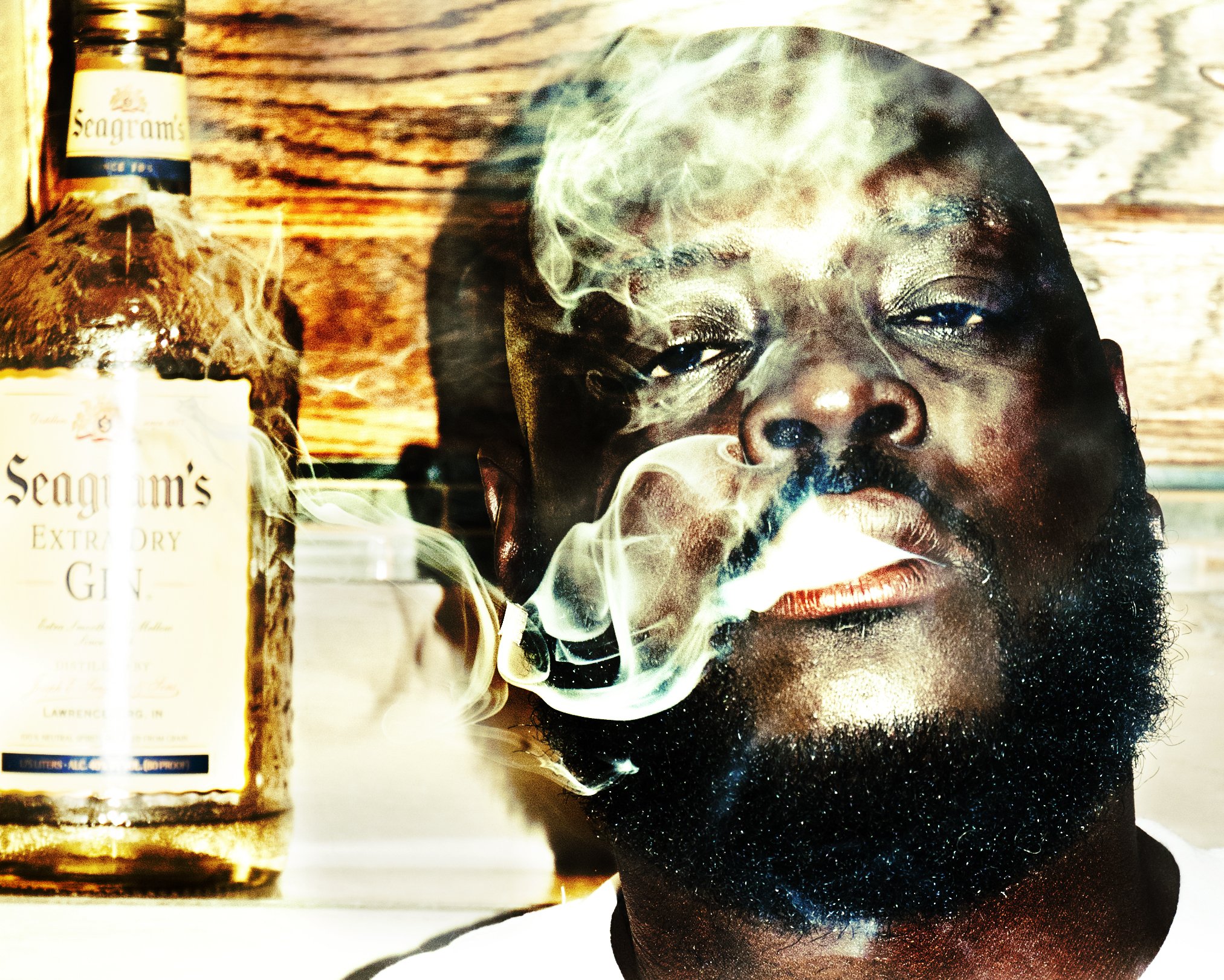 A close-up of a man’s face with dark skin, a beard, and closed eyes, smoking a cigarette with smoke swirling around his face. On the left side, there are bottles of Seagram's gin, with one in the foreground and another behind it, placed on a wooden surface.