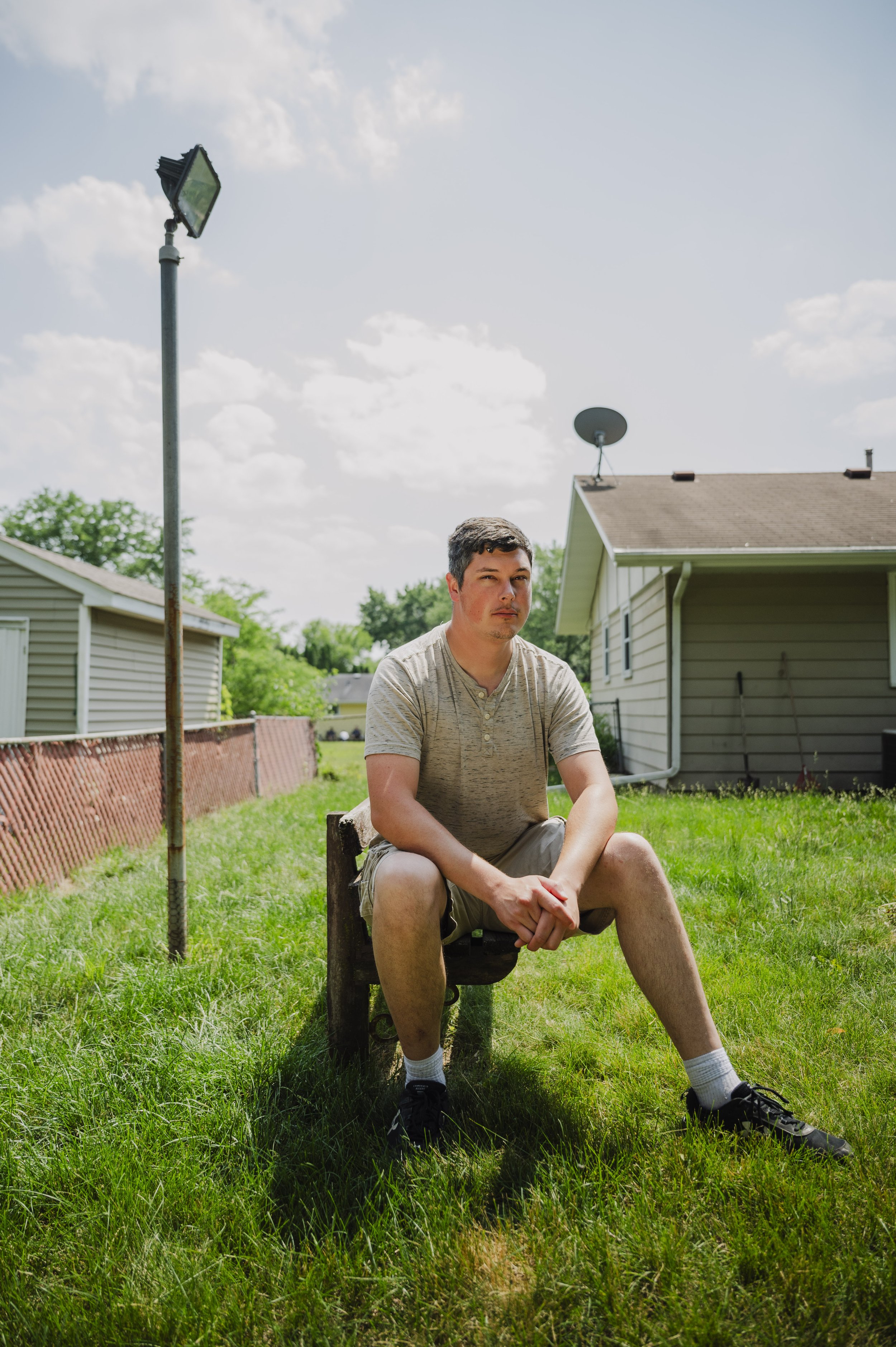 A young man sitting outdoors on a small wooden bench in a grassy backyard, with a light gray house, satellite dish, and a tall floodlight in the background on a partly cloudy day.