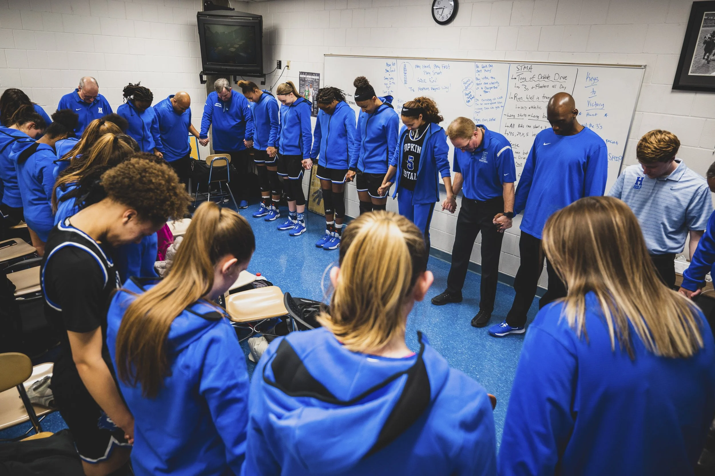 A group of young athletes, dressed in blue sportswear, standing in a circle holding hands inside a classroom or meeting room, likely participating in a prayer or moment of silence.