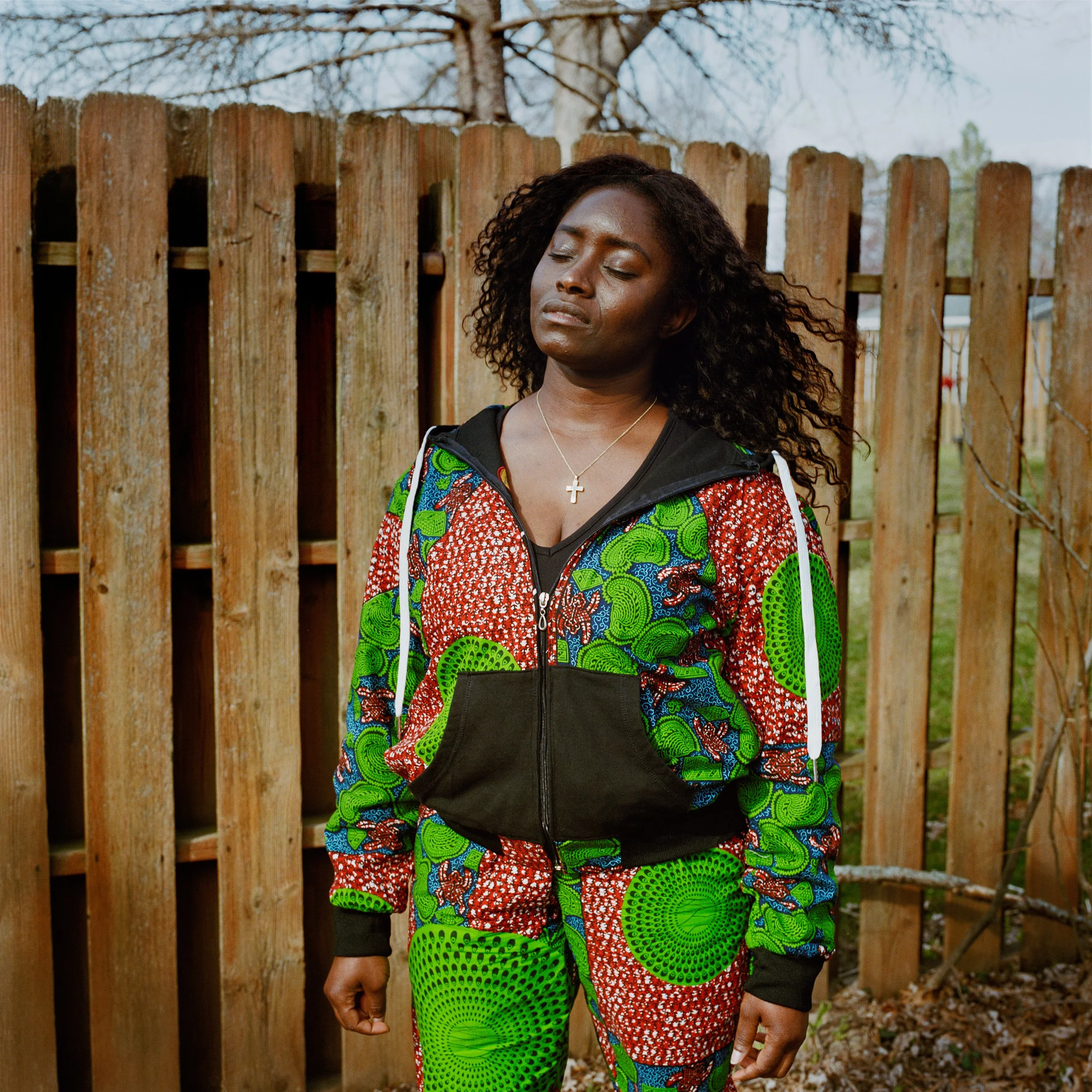 A Black woman with closed eyes and a somber expression standing outdoors in front of a wooden fence, wearing a colorful, patterned tracksuit and a necklace with a cross. She is contemplating the death of her mother from COVID-19.