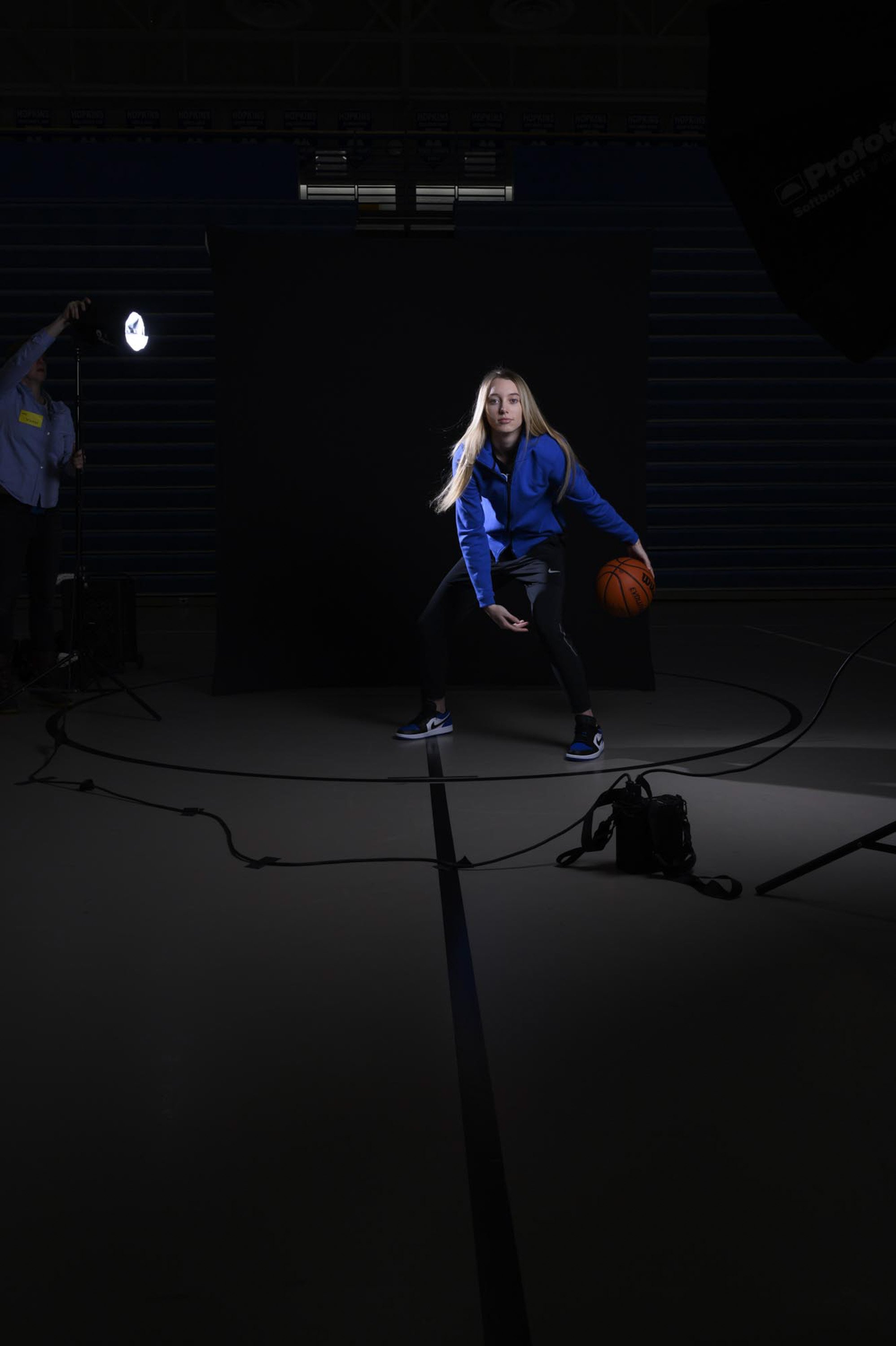 A woman in sportswear is posing with a basketball on a dark indoor basketball court with professional lighting equipment around her.