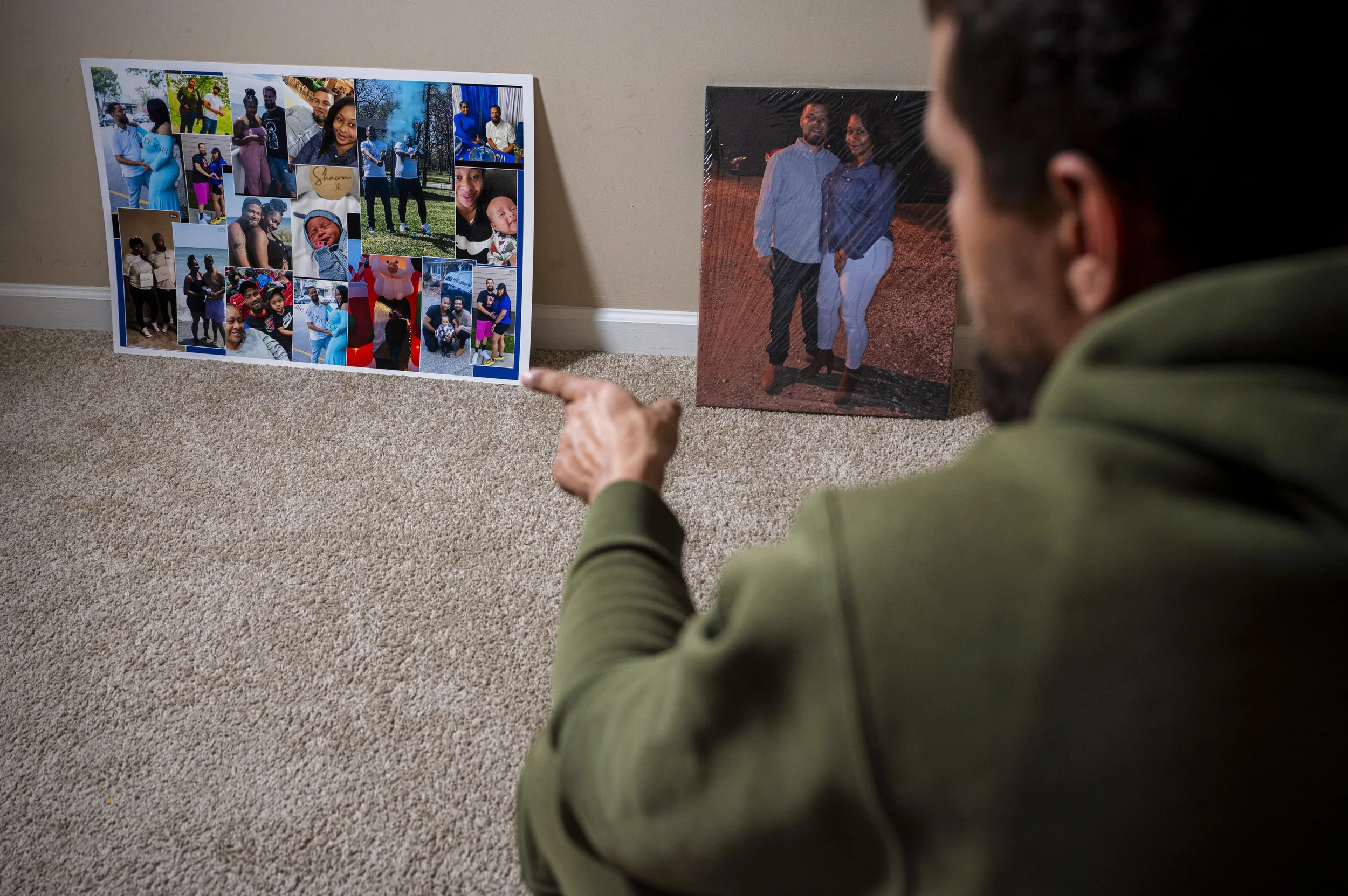 A man with dark hair points at a collage of various photographs on a white poster board placed on the carpeted floor. Next to the collage, a large framed photograph of a couple walking outdoors at night is visible.